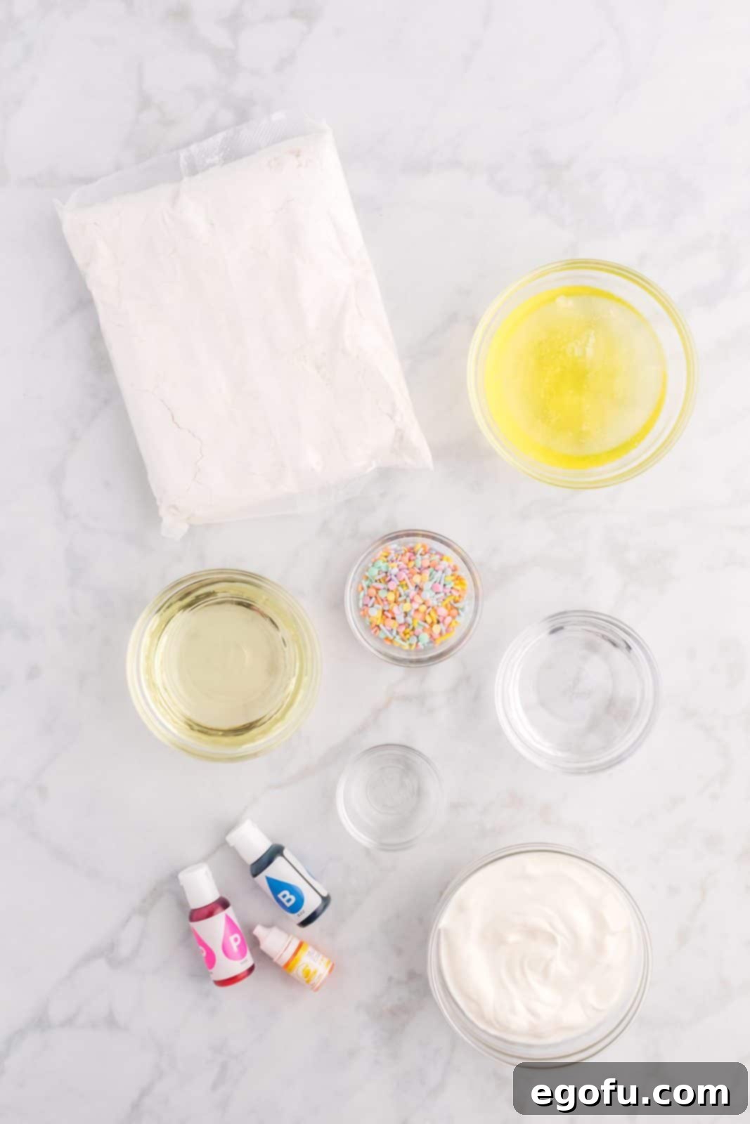 Arrangement of all ingredients needed: white cake mix, water, egg whites, vegetable oil, sour cream, clear vanilla extract, blue, pink, and yellow food coloring, pastel colored sprinkles, softened butter, powdered sugar, salt, and heavy cream.