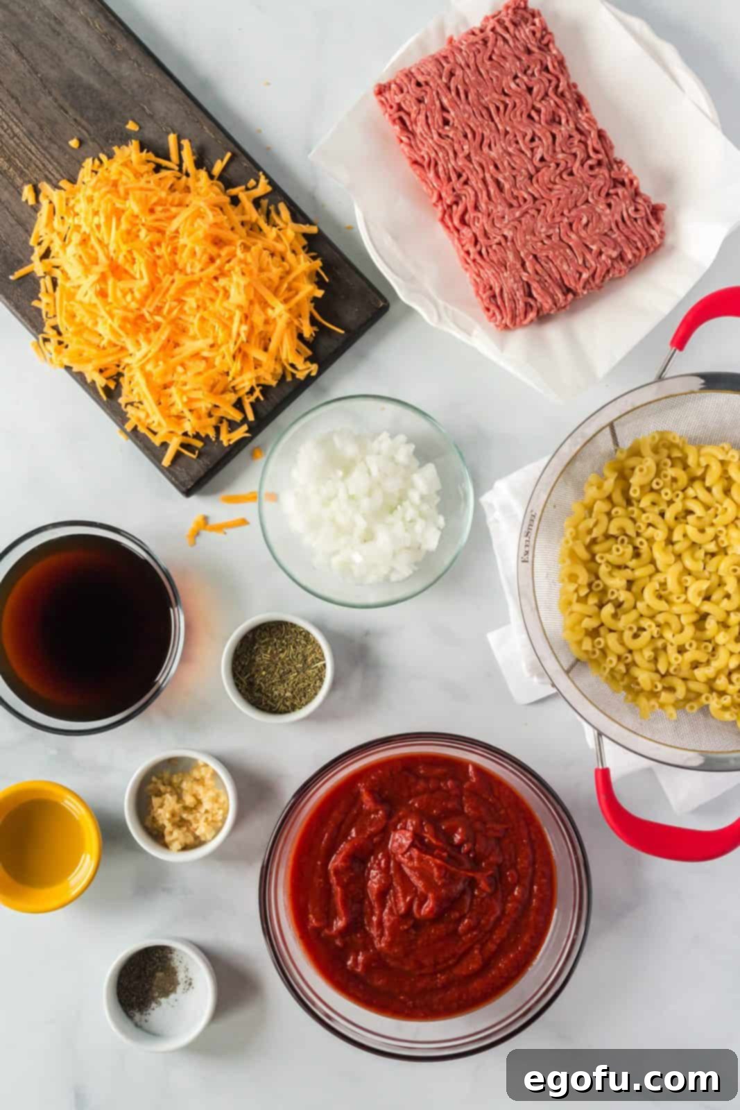 Close-up shot of all the individual ingredients laid out, including diced white onion, a bottle of olive oil, a shaker of Italian seasoning, minced garlic, salt, pepper, a package of ground beef, dry elbow macaroni, a can of crushed tomatoes, a container of beef broth, a bag of shredded cheddar cheese, and fresh parsley sprigs.