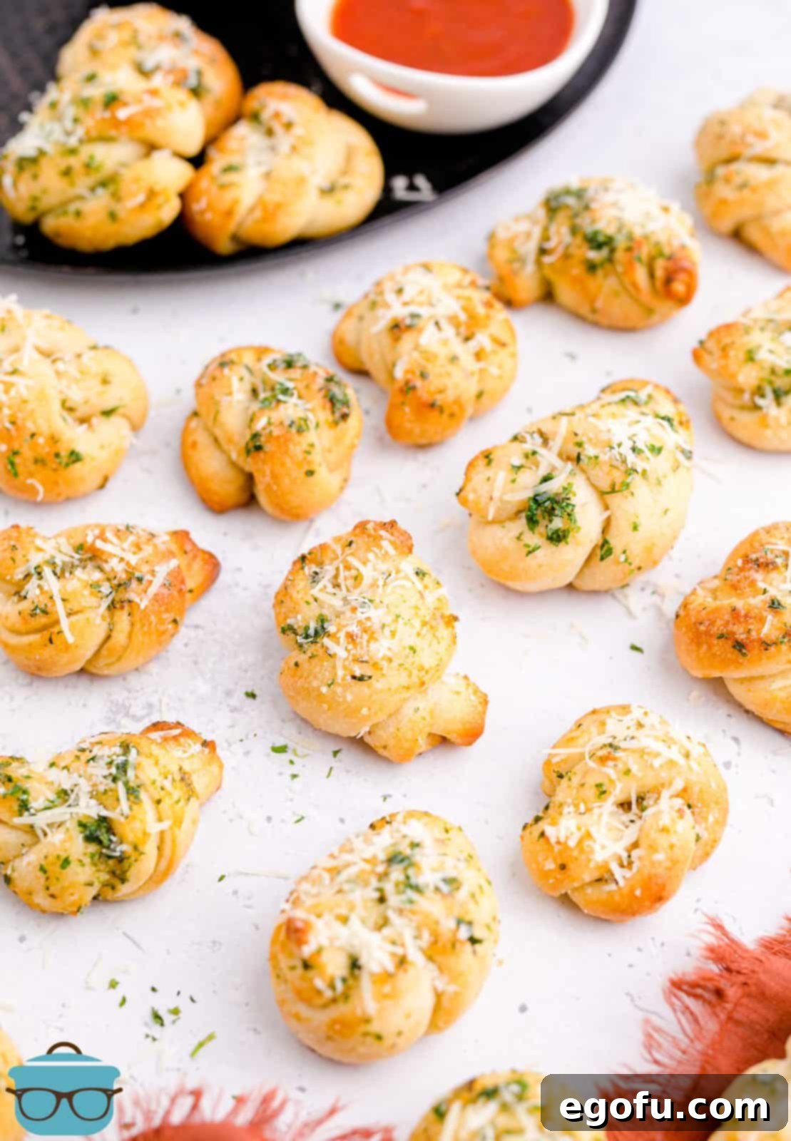 Finished Easy Garlic Knots piled on a white board, lightly sprinkled with cheese, next to a bowl of marinara sauce.