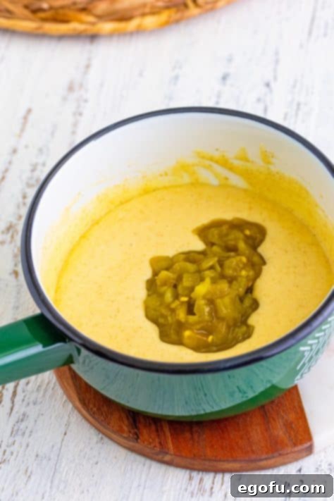 Green chiles being added to the creamy Queso Dip in a saucepan.