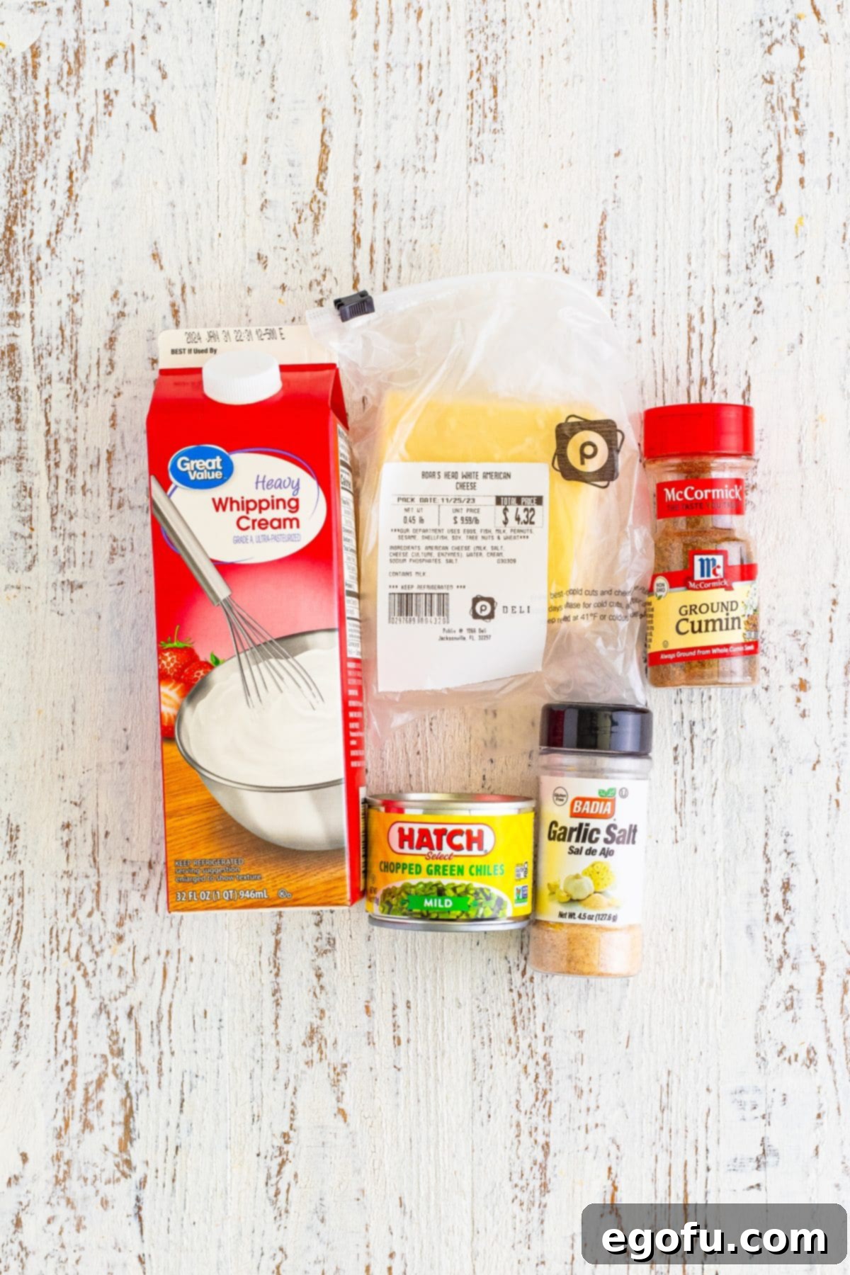 The five main ingredients for Queso Dip laid out: a carton of heavy cream, a block of white American cheese, ground cumin, garlic salt, and a can of chopped green chiles.