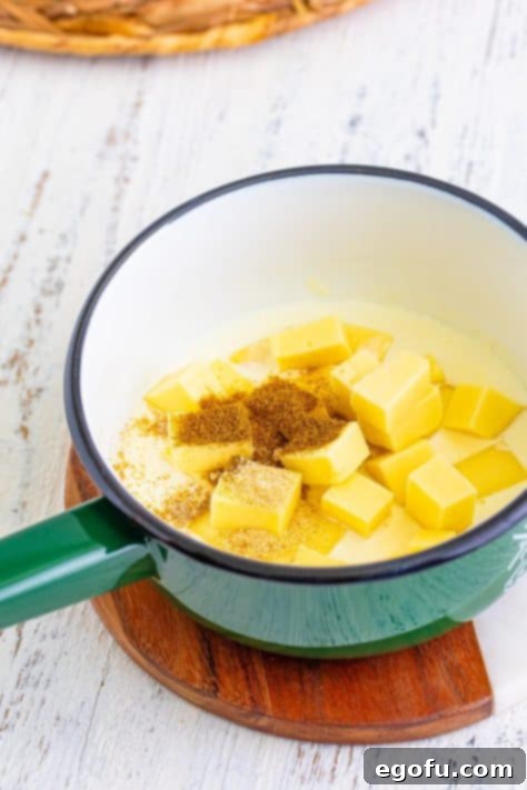 Blocks of cheese, cumin, and garlic salt being added to heated heavy cream in a pot.