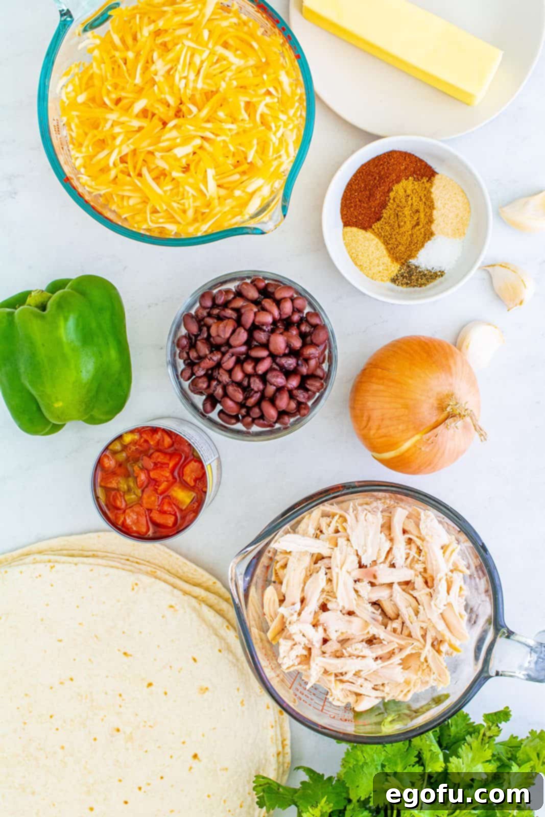 A vibrant display of ingredients for Sheet Pan Chicken Quesadillas: fresh onions, green bell peppers, garlic, assorted spices, shredded chicken, canned black beans, Rotel, fresh cilantro, large tortillas, and shredded Colby Jack cheese.