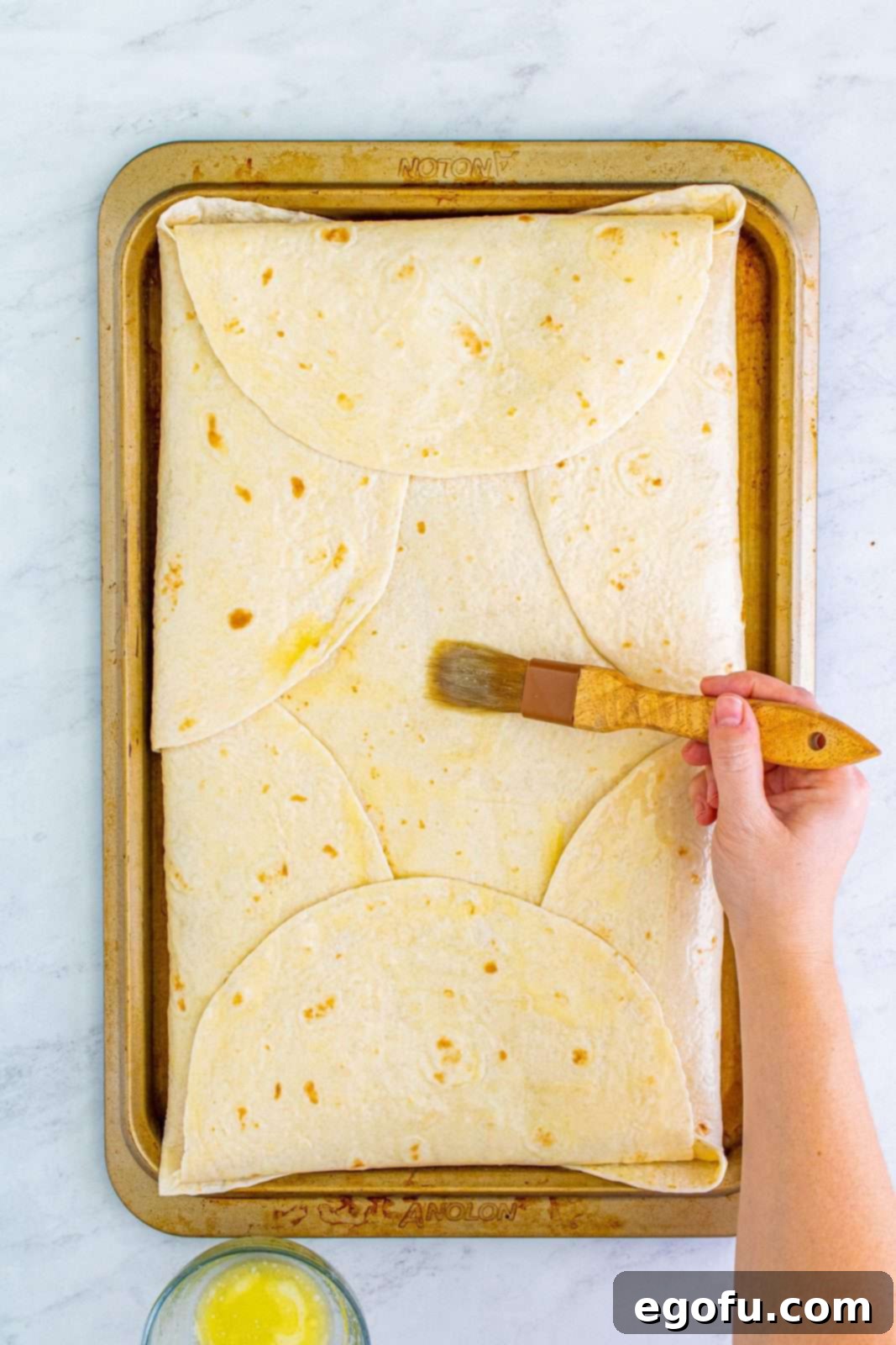 Melted butter being brushed generously over the neatly folded edges of the large quesadilla on the sheet pan, preparing it for baking.