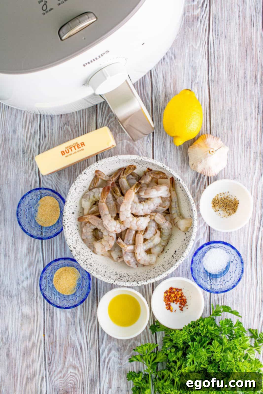 A flat lay photograph showcasing all the fresh and dry ingredients required for Air Fryer Garlic Butter Shrimp, neatly arranged: large shrimp, olive oil, kosher salt, garlic powder, onion powder, black pepper, olive oil spray, unsalted butter, fresh garlic, lemon juice, red pepper flakes, and fresh parsley.