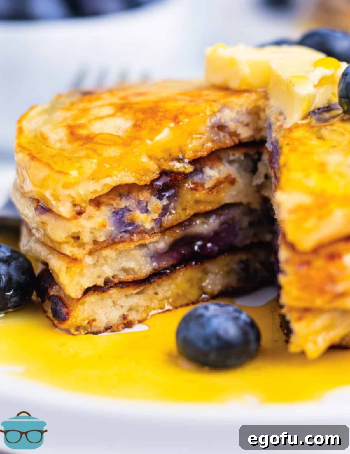 A towering stack of golden-brown fluffy blueberry pancakes, glistening with maple syrup and topped with fresh blueberries and a dollop of butter.