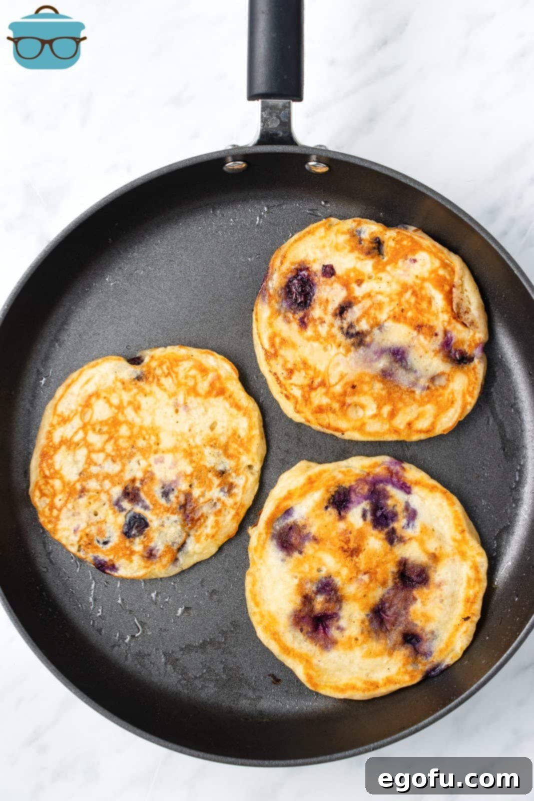 Several blueberry pancakes cooking on a hot frying pan, showing bubbles forming on the surface.