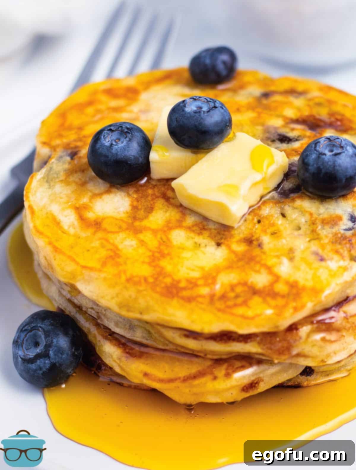 A stack of perfectly cooked blueberry pancakes on a white plate, adorned with butter, maple syrup, and fresh blueberries, ready to be served.