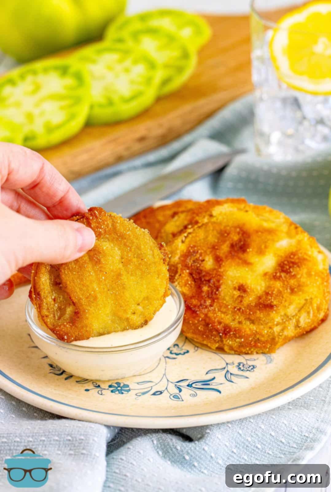 A hand gracefully dips a golden-brown Fried Green Tomato into a creamy white dipping sauce, showcasing the perfect way to enjoy this classic Southern delicacy.