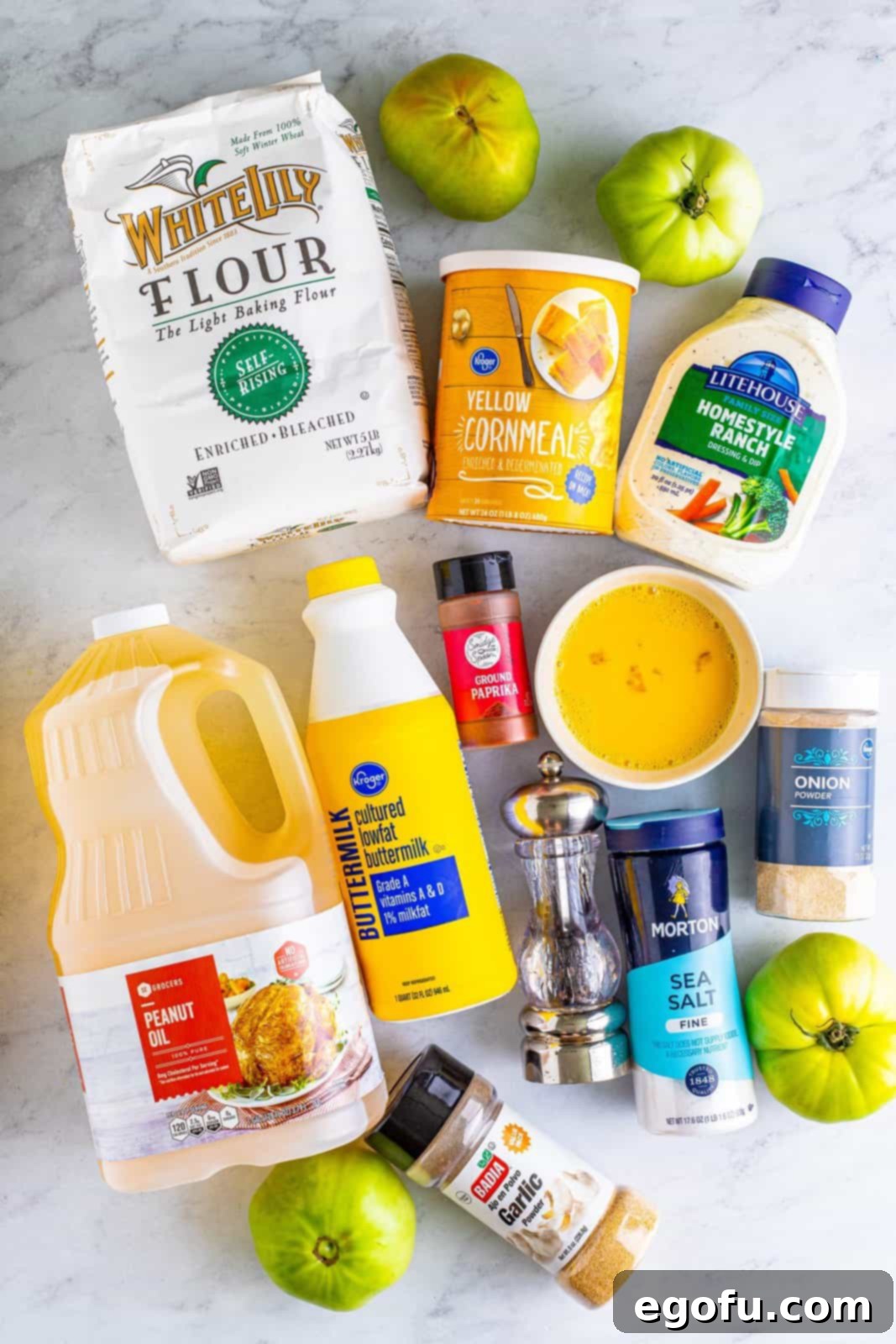 An organized display of fresh ingredients for Fried Green Tomatoes: self-rising flour, creamy buttermilk, eggs, black pepper, salt, garlic powder, onion powder, sweet paprika, vibrant yellow cornmeal, firm green tomatoes, a bottle of peanut oil, and a small bowl of ranch dressing for serving.