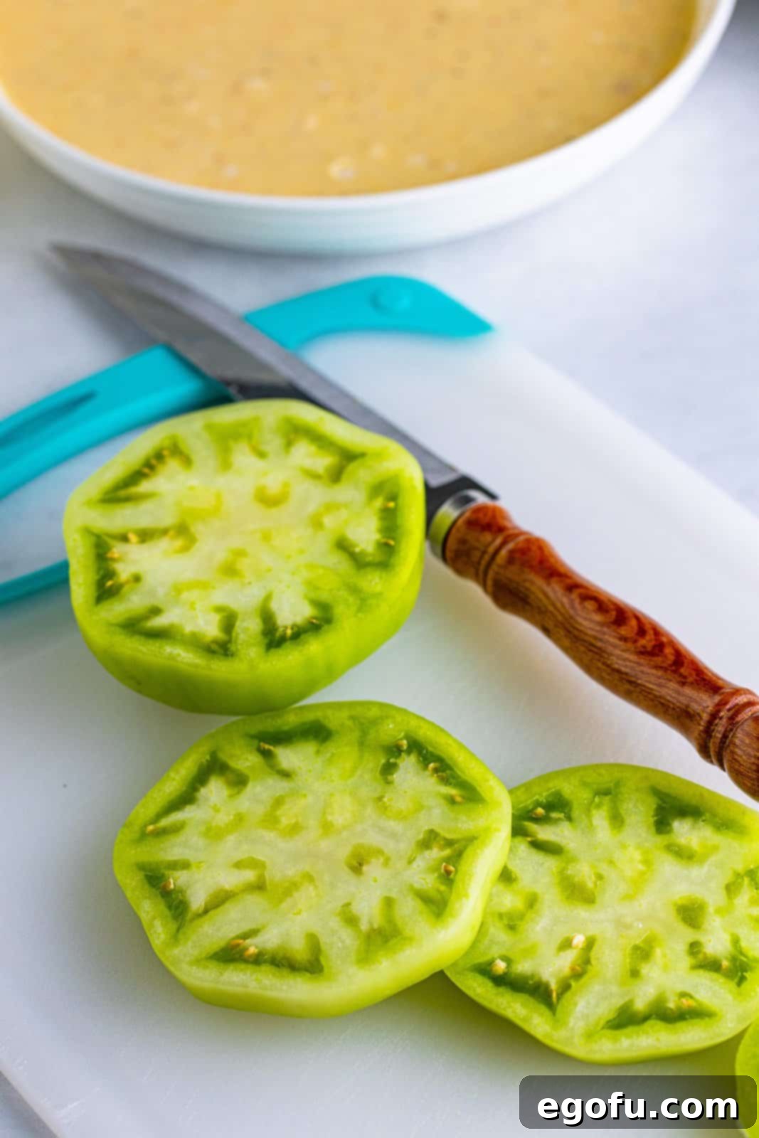 A sharp knife slices a firm green tomato into uniform ¼-inch thick rounds, preparing them for the classic Southern fried green tomato recipe.