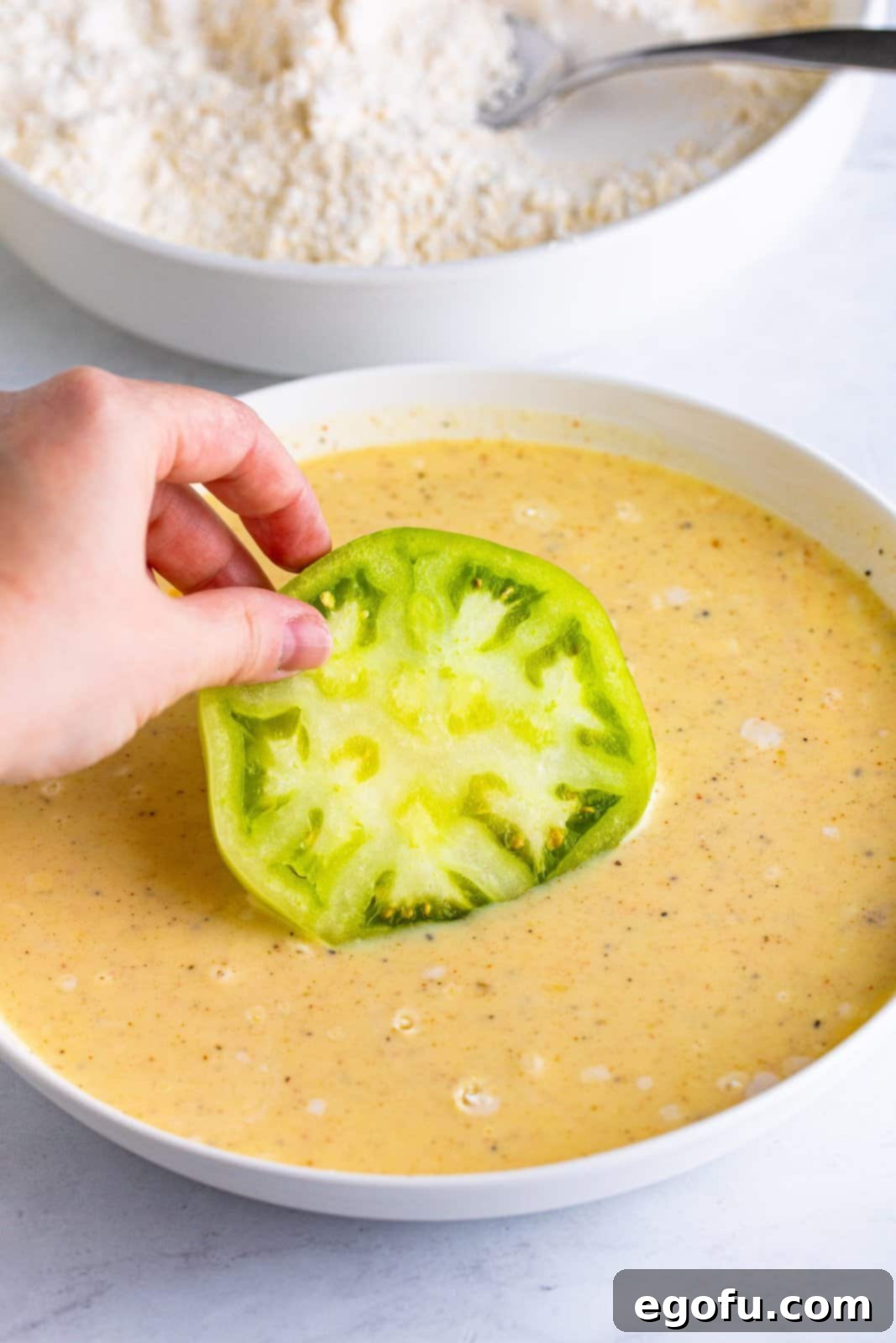 A thick slice of green tomato is gently dipped and coated in the creamy buttermilk mixture, the first step in creating crispy fried green tomatoes.