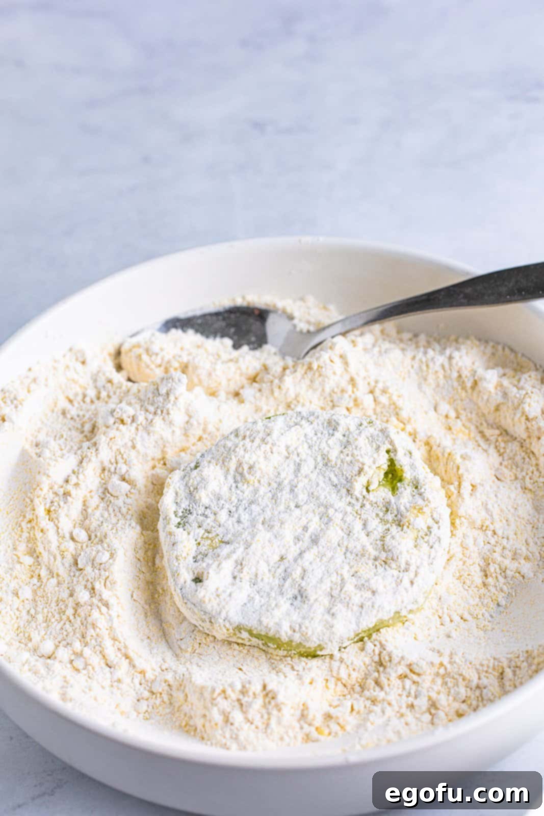 A buttermilk-coated green tomato slice is carefully transferred into the flour and cornmeal mixture, ensuring an even and thorough dry coating for frying.