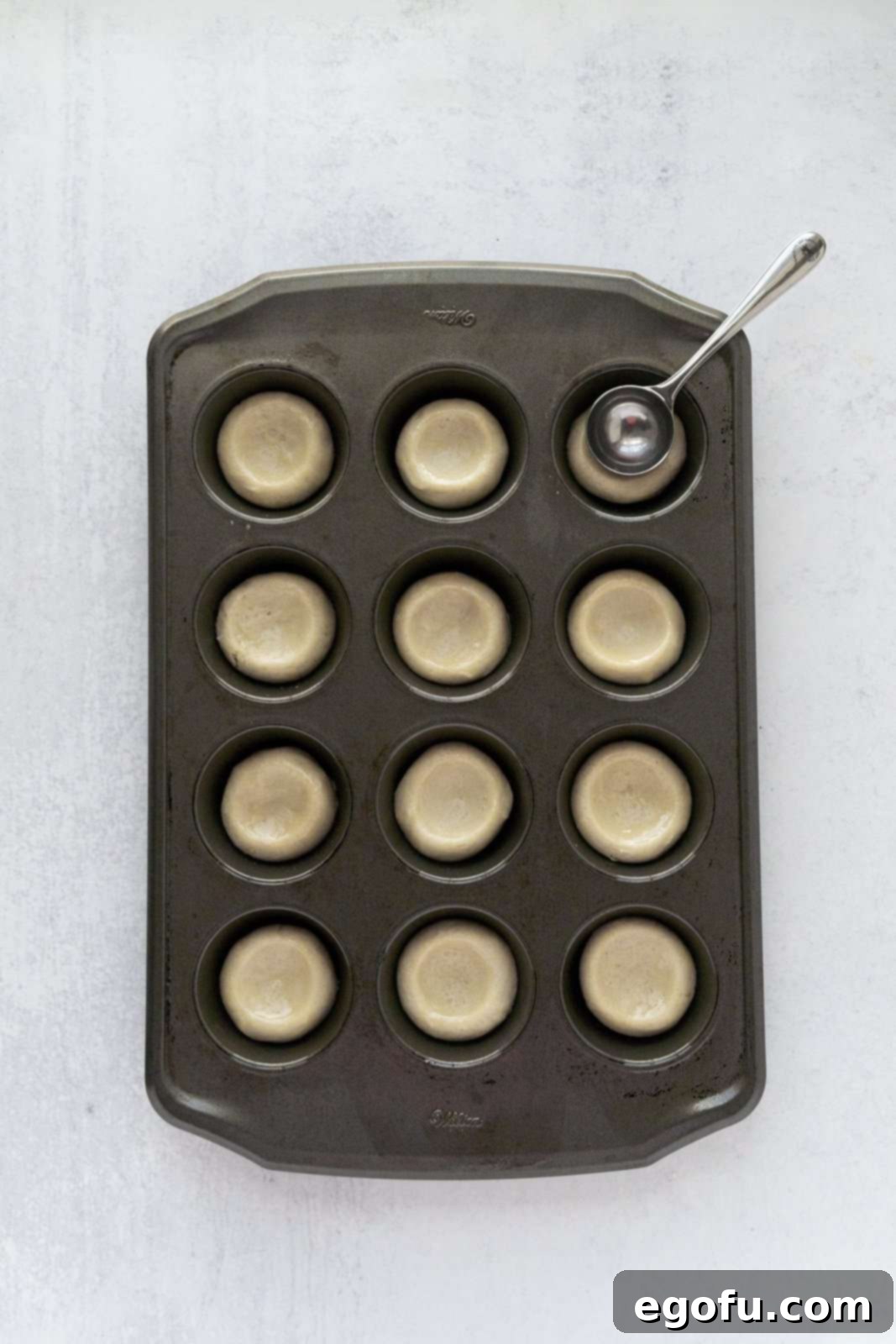 Cookie dough added to mini muffin tin with measuring spoon pressing down depressions in dough.