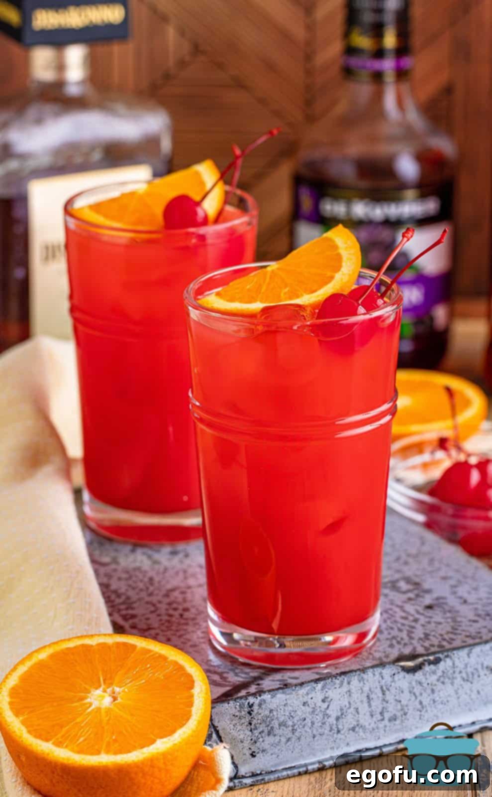 Dixie Dynamite 2 Two Alabama Slammer cocktails in glasses, garnished with orange slices and maraschino cherries, resting on a metal tray with alcohol bottles blurred in the background.