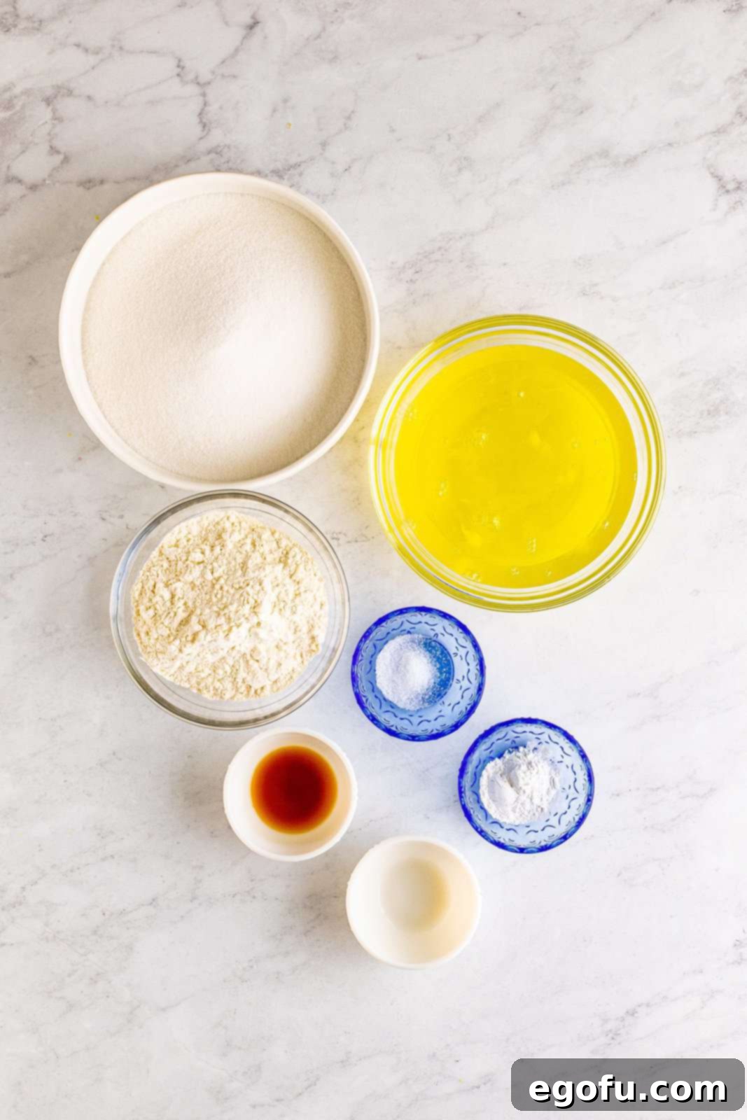 Ingredients needed for Angel Food Cake laid out on a table: granulated sugar, all-purpose flour, fine sea salt, egg whites, cream of tartar, vanilla extract, and almond extract.