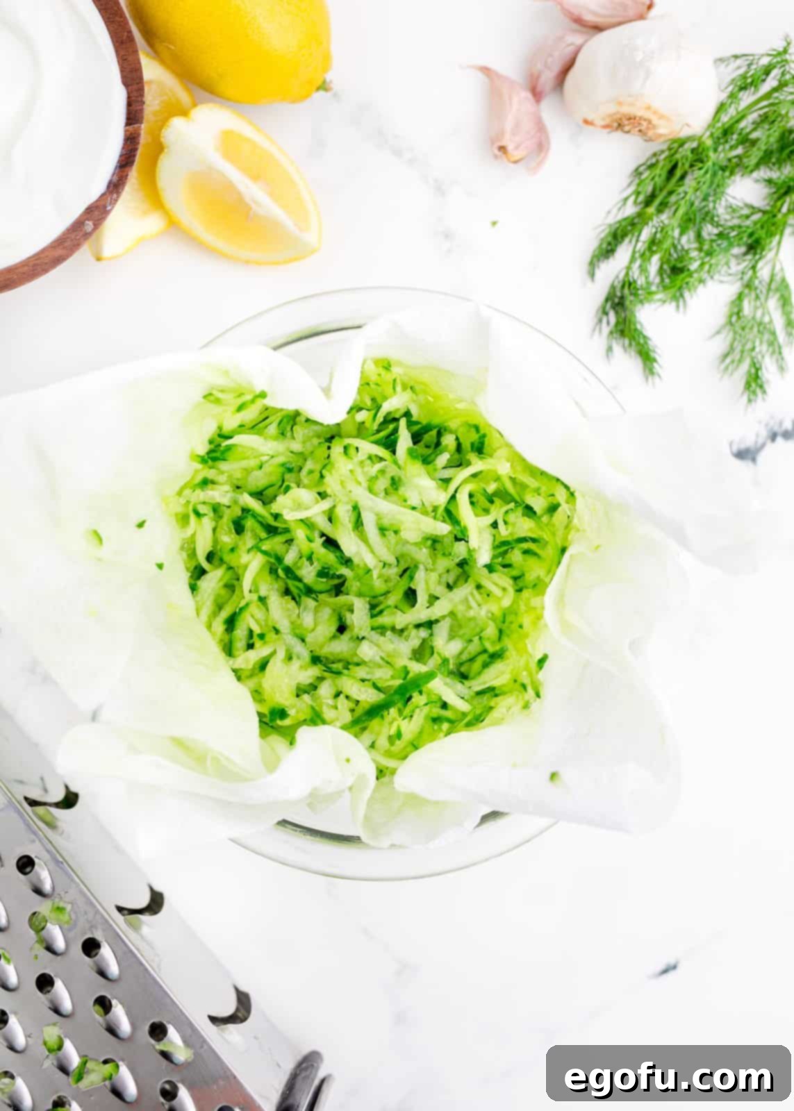 Grated cucumber being drained thoroughly in paper towels over a bowl.