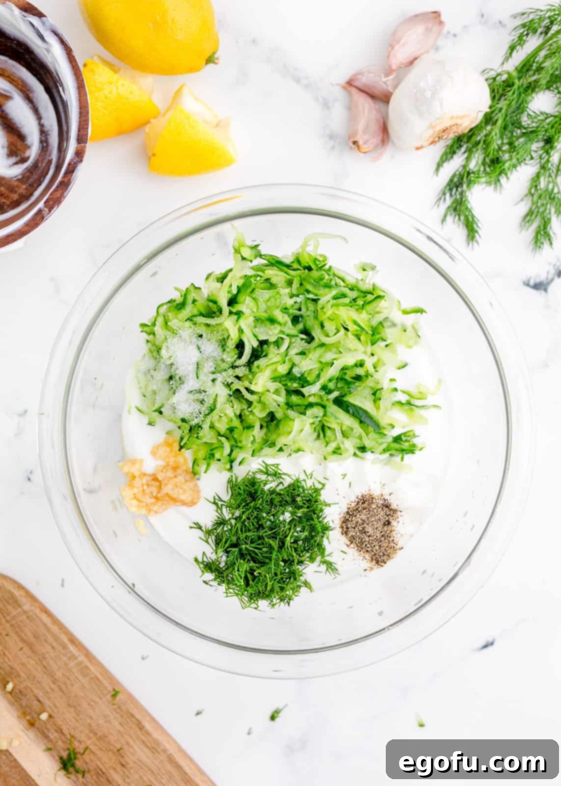 Tzatziki sauce ingredients added to a bowl, showing yogurt, drained cucumber, dill, lemon juice, olive oil, and salt.