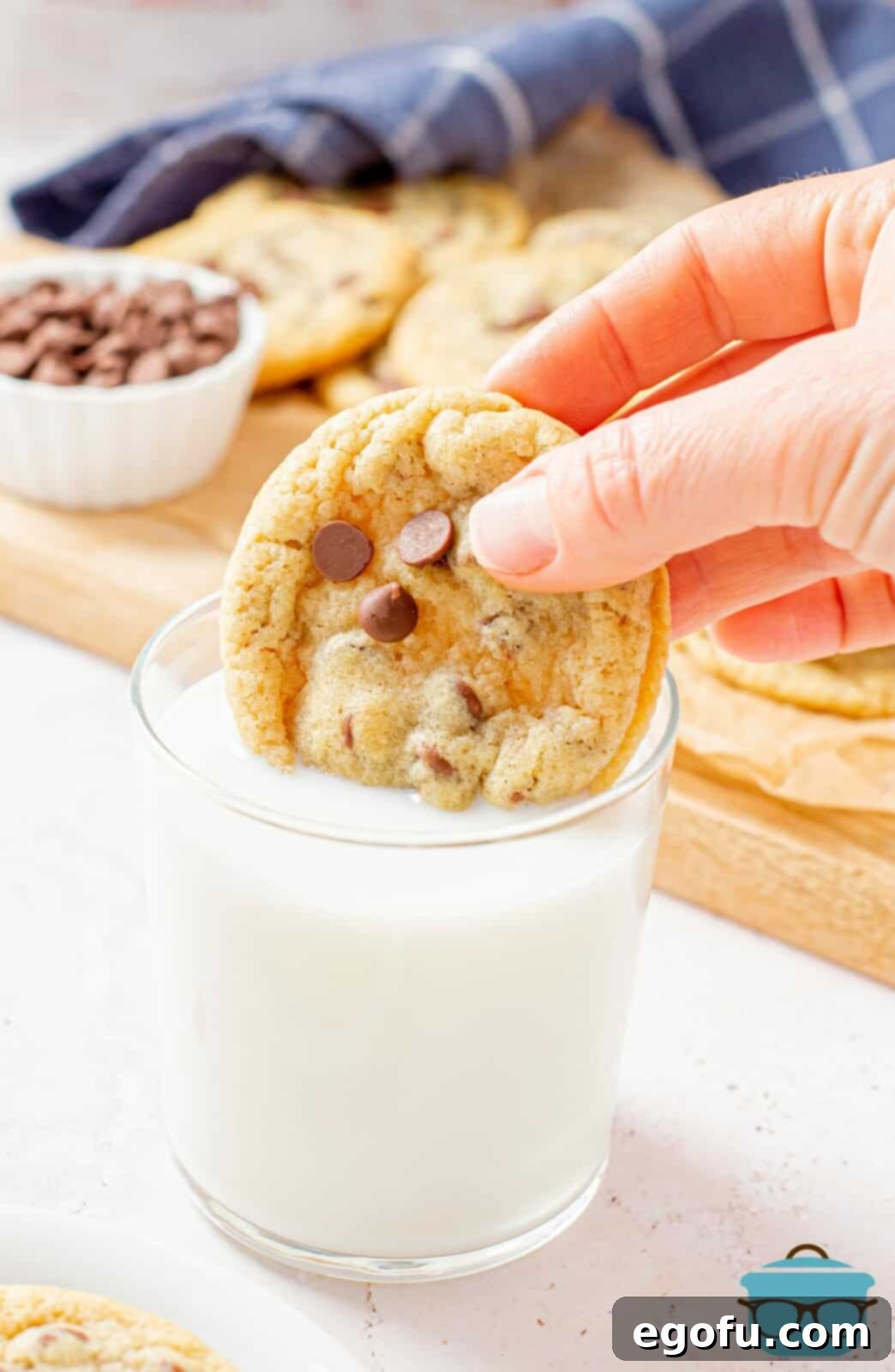 A hand gently dipping a warm chewy chocolate chip cookie into a glass of cold milk.