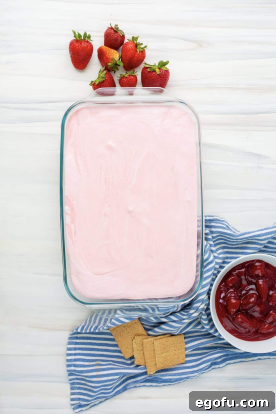 The vibrant strawberry pudding mixture being poured and spread evenly over the second layer of graham crackers in the dish.