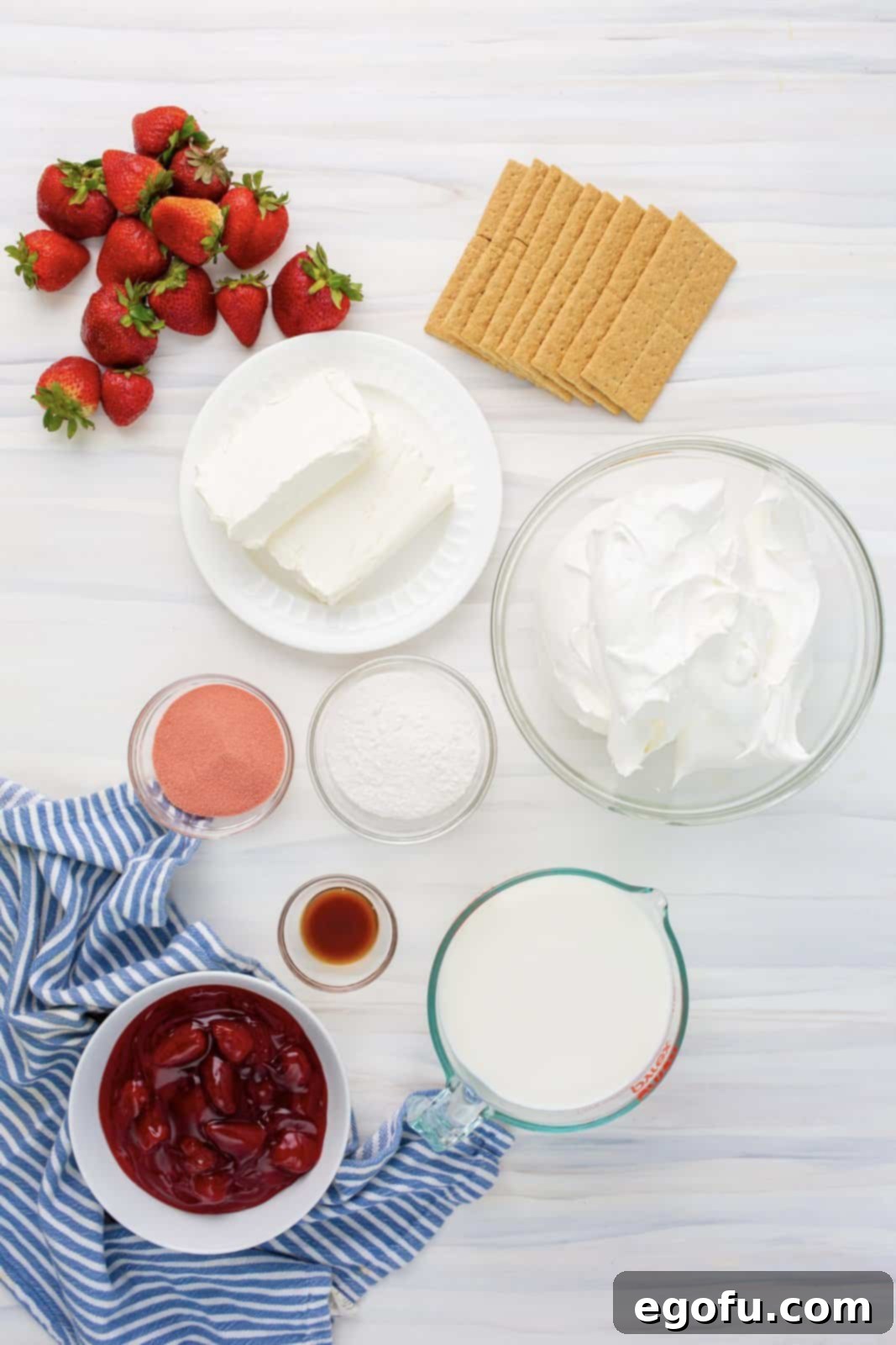 A flat lay photograph showcasing all the ingredients required for making the Strawberry Icebox Cake: cream cheese, instant cheesecake pudding mix, strawberry Jell-O mix, fresh strawberries, Cool Whip, milk, vanilla extract, graham crackers, and optional strawberry pie filling.