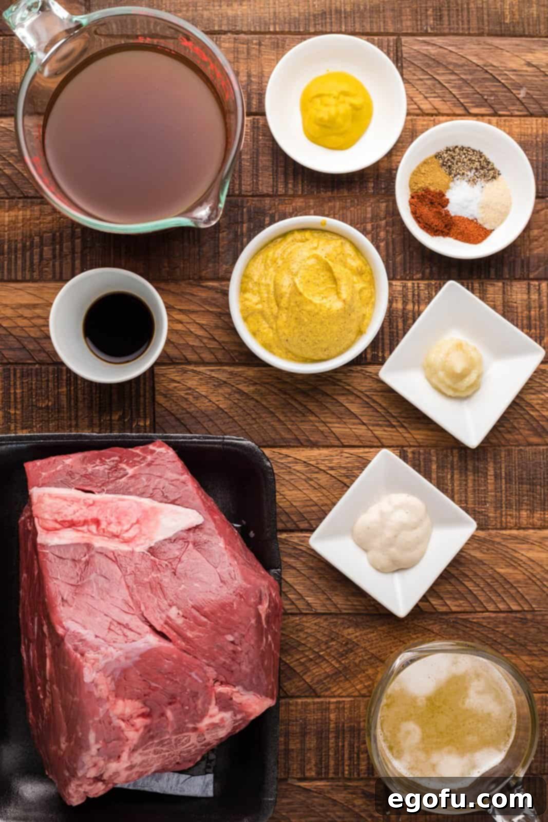 Pitmaster's Smoked Rump 4 A collection of ingredients laid out on a kitchen counter: a rump roast, containers of beef broth and pale lager beer, bowls of brown mustard, garlic paste, horseradish, yellow mustard, Worcestershire sauce, salt, pepper, paprika, cumin, chili powder, and onion powder.