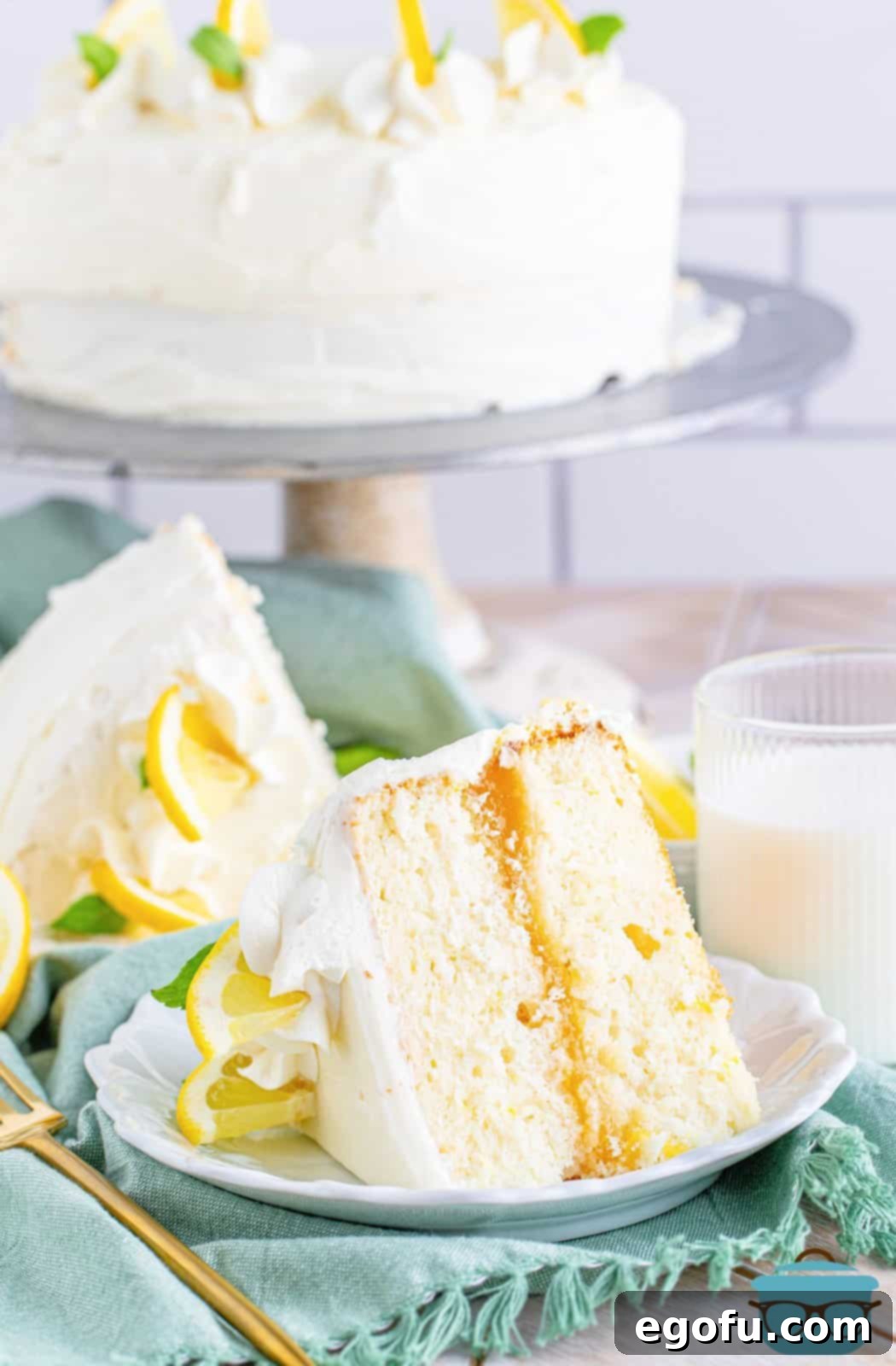 Zesty Lemon Dream Cake 2 Slice of Lemon Layer Cake on white plate showing filling with whole cake in background on stand.
