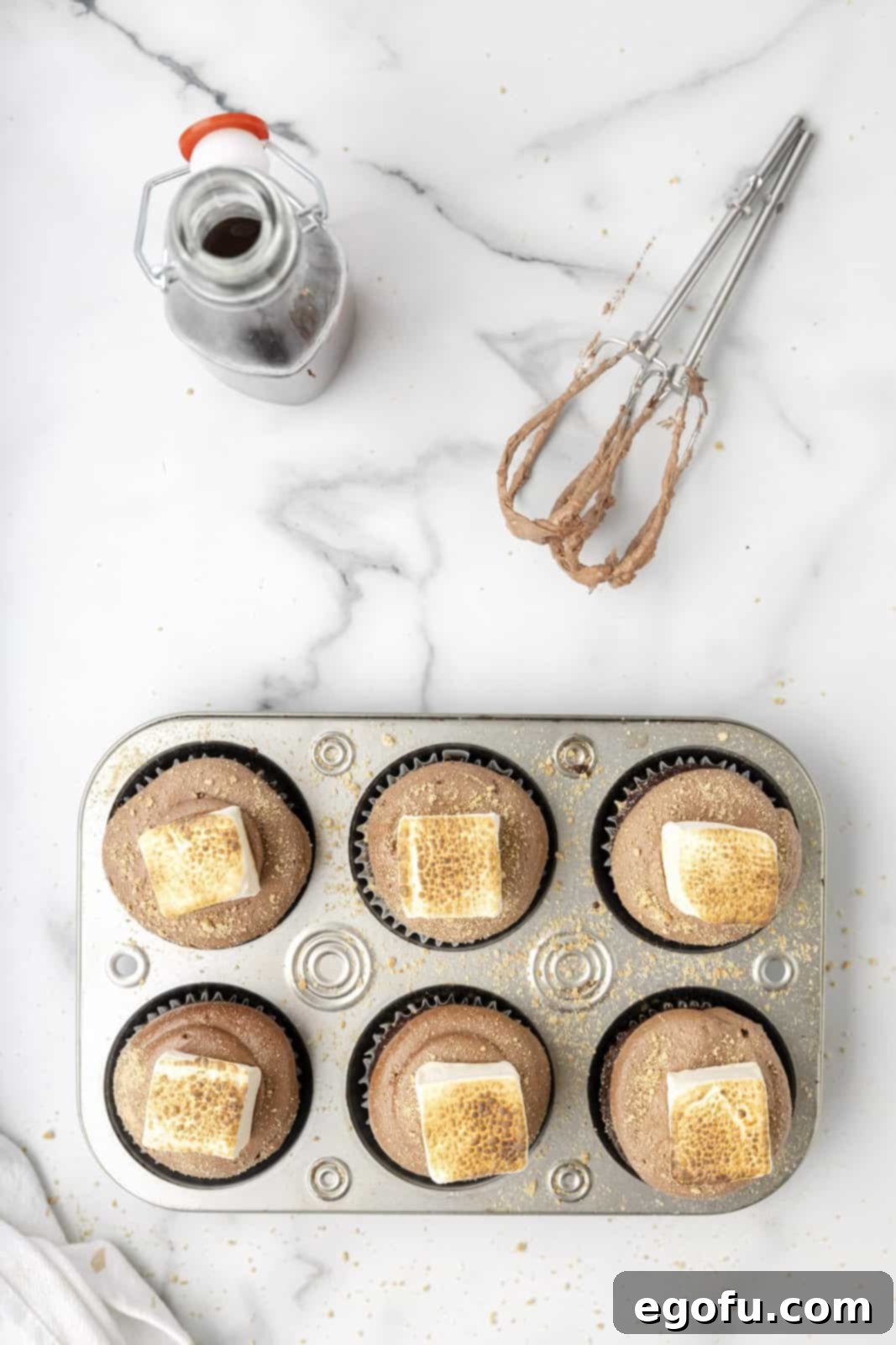 A perfectly toasted marshmallow is gently placed on top of a frosted cupcake, completing the S'mores creation.