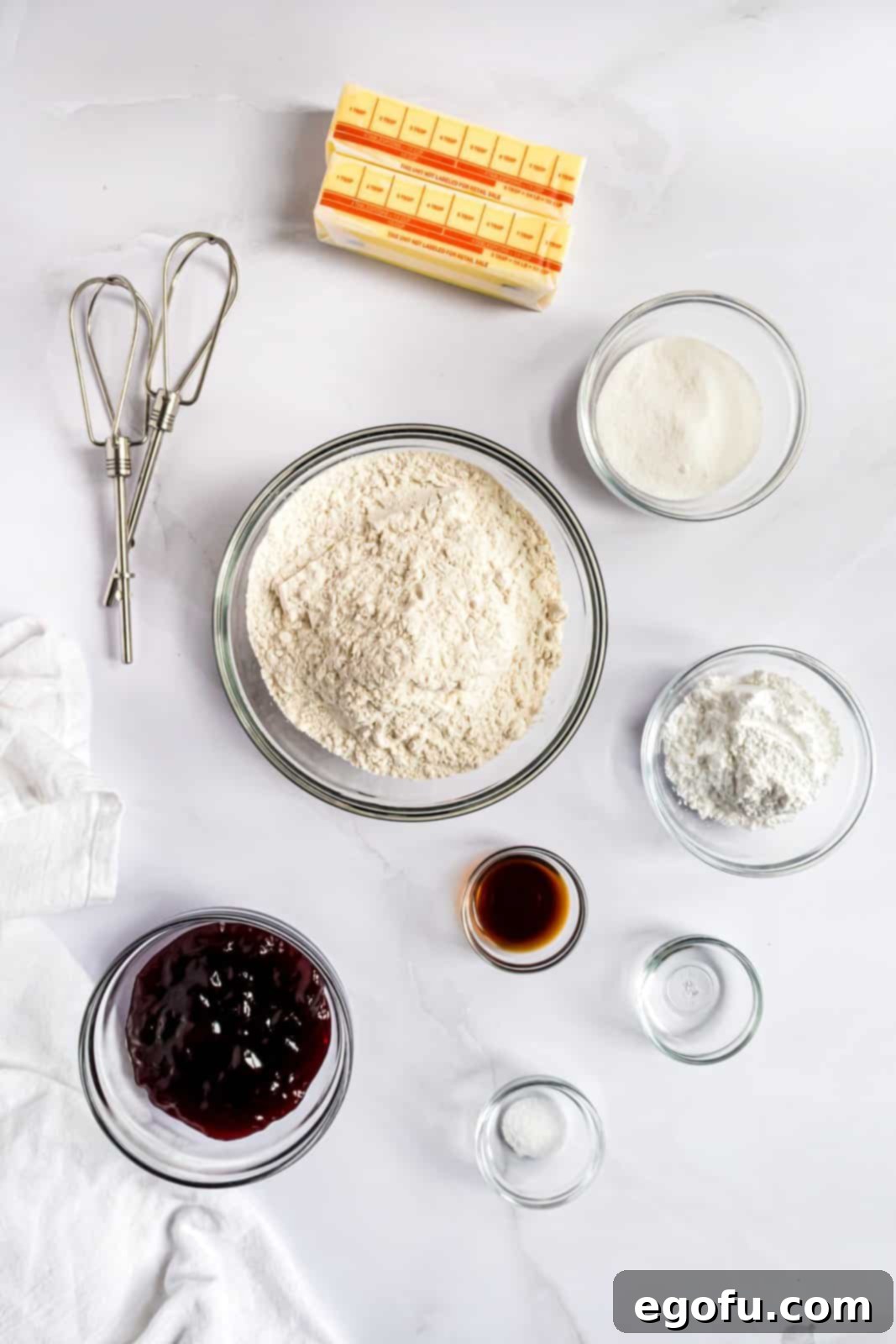 A collection of ingredients artfully arranged for making Raspberry Tartlets, including softened unsalted butter, granulated sugar, powdered sugar, vanilla extract, almond extract, all-purpose flour, kosher salt, and a jar of seedless raspberry jam.