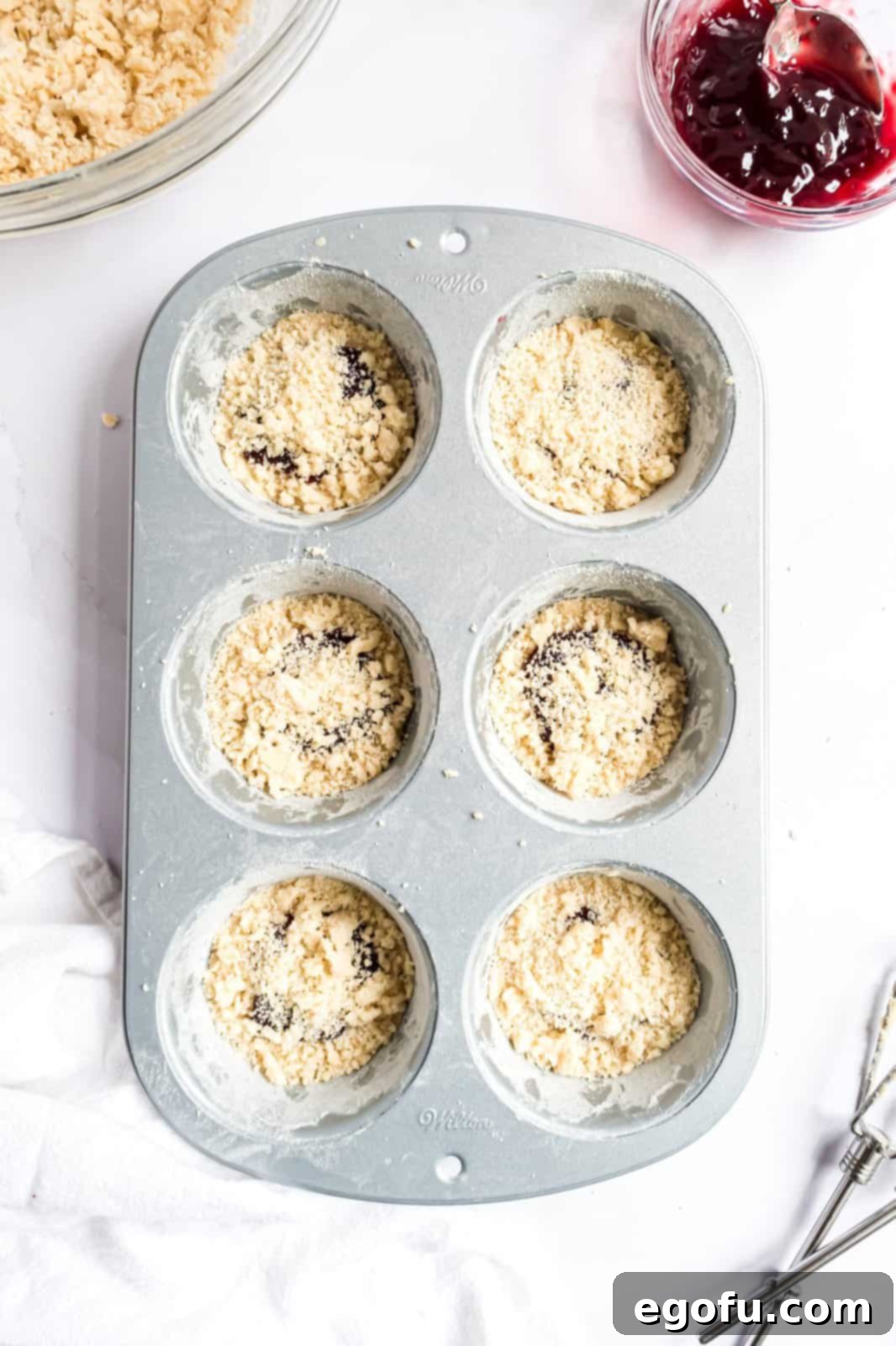 The delicate crust crumbles are generously sprinkled over the luscious raspberry jam, creating an inviting topping for the tartlets in the tin.