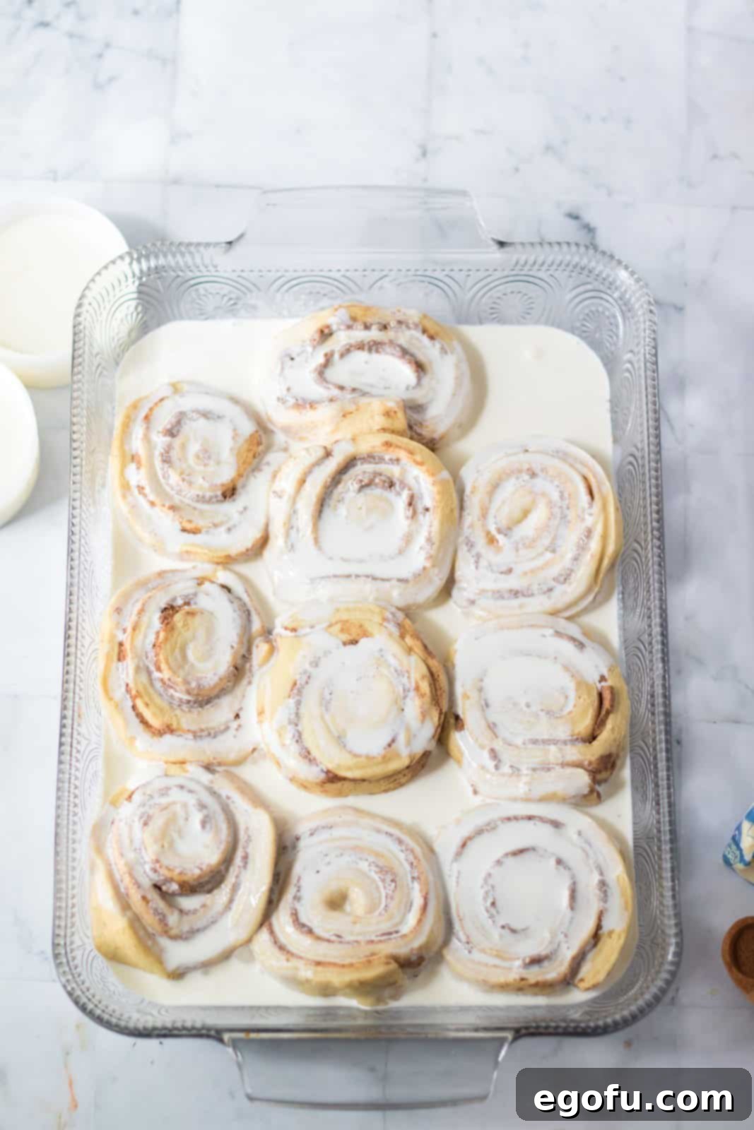 Raw cinnamon rolls arranged in a baking dish, with heavy cream being poured over them.