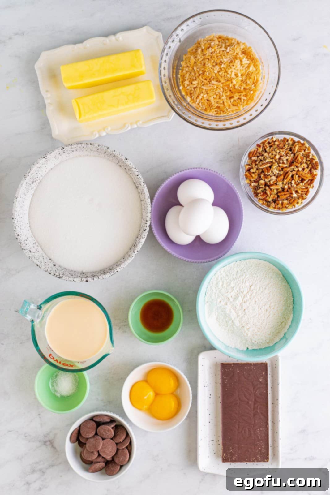 A selection of fresh ingredients laid out, including salted butter, German chocolate baking bar, granulated sugar, eggs, vanilla extract, fine sea salt, all-purpose flour, evaporated milk, egg yolks, toasted coconut, chopped pecans, and chocolate melting wafers.
