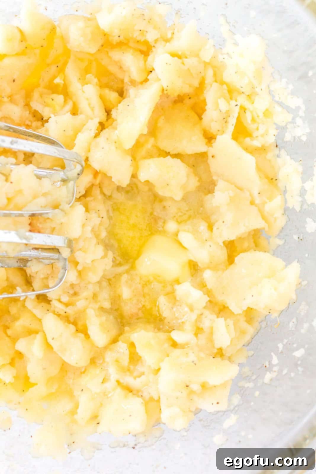 Potato mixture with seasonings, chicken broth, and butter being blended in a bowl using a hand mixer, showing a partially combined texture.