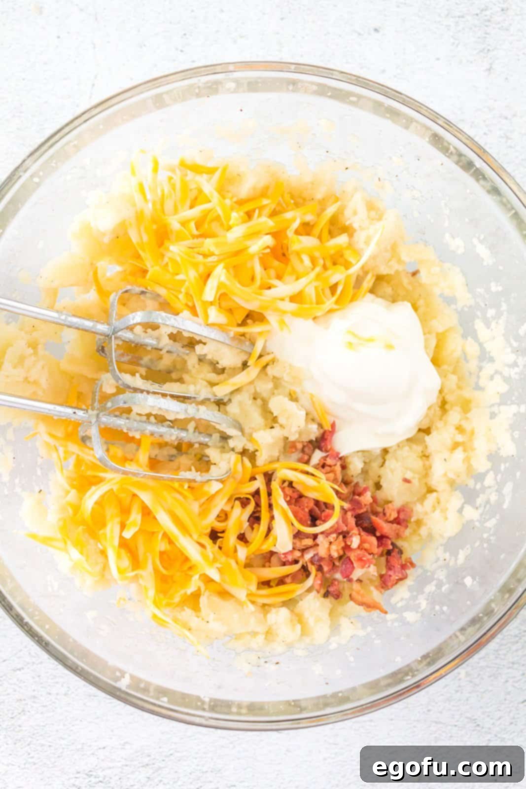 Sour cream, bacon bits, and shredded Colby Jack cheese added to the potato mixture in a bowl, ready for the final blend.