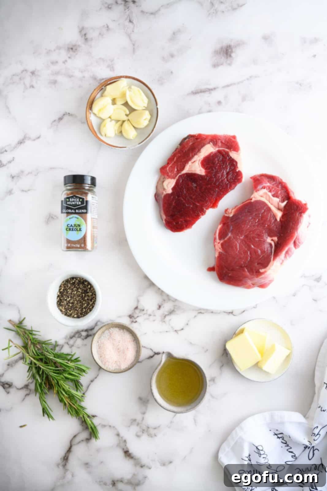 Ingredients needed for pan-seared steak: rib-eye steaks, salt, pepper, cajun creole seasoning, olive oil, unsalted butter, garlic cloves and rosemary sprigs displayed on a cutting board.