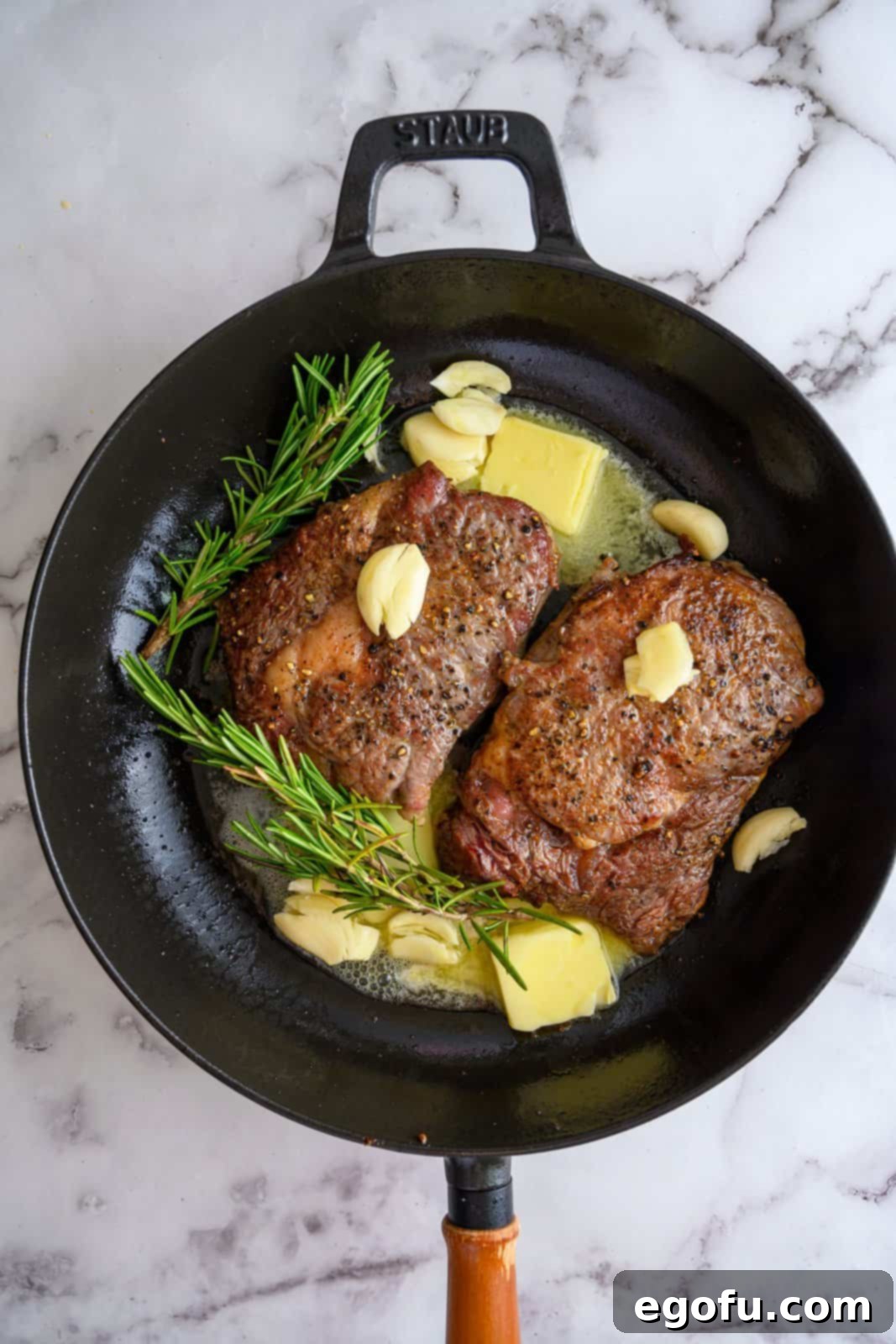 Steaks searing in a cast iron pan with butter, smashed garlic cloves, and fresh rosemary sprigs.
