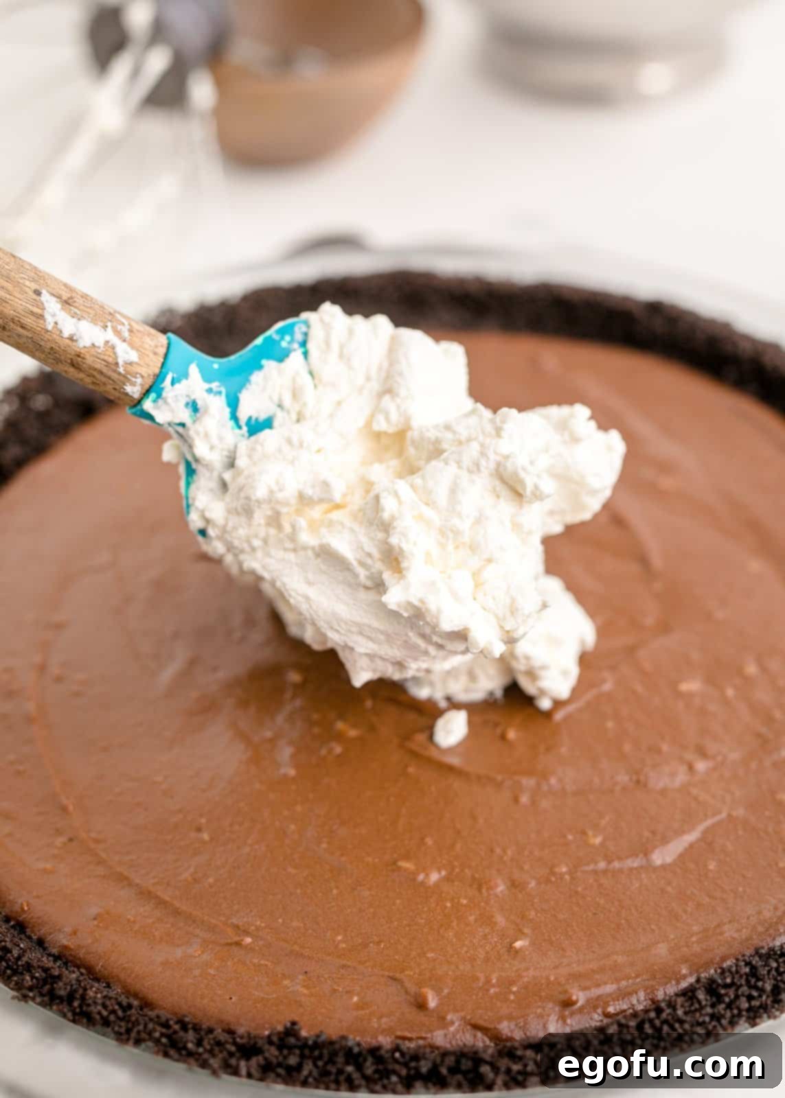 Whipped cream being spread evenly on top of the chilled chocolate cream pie filling.