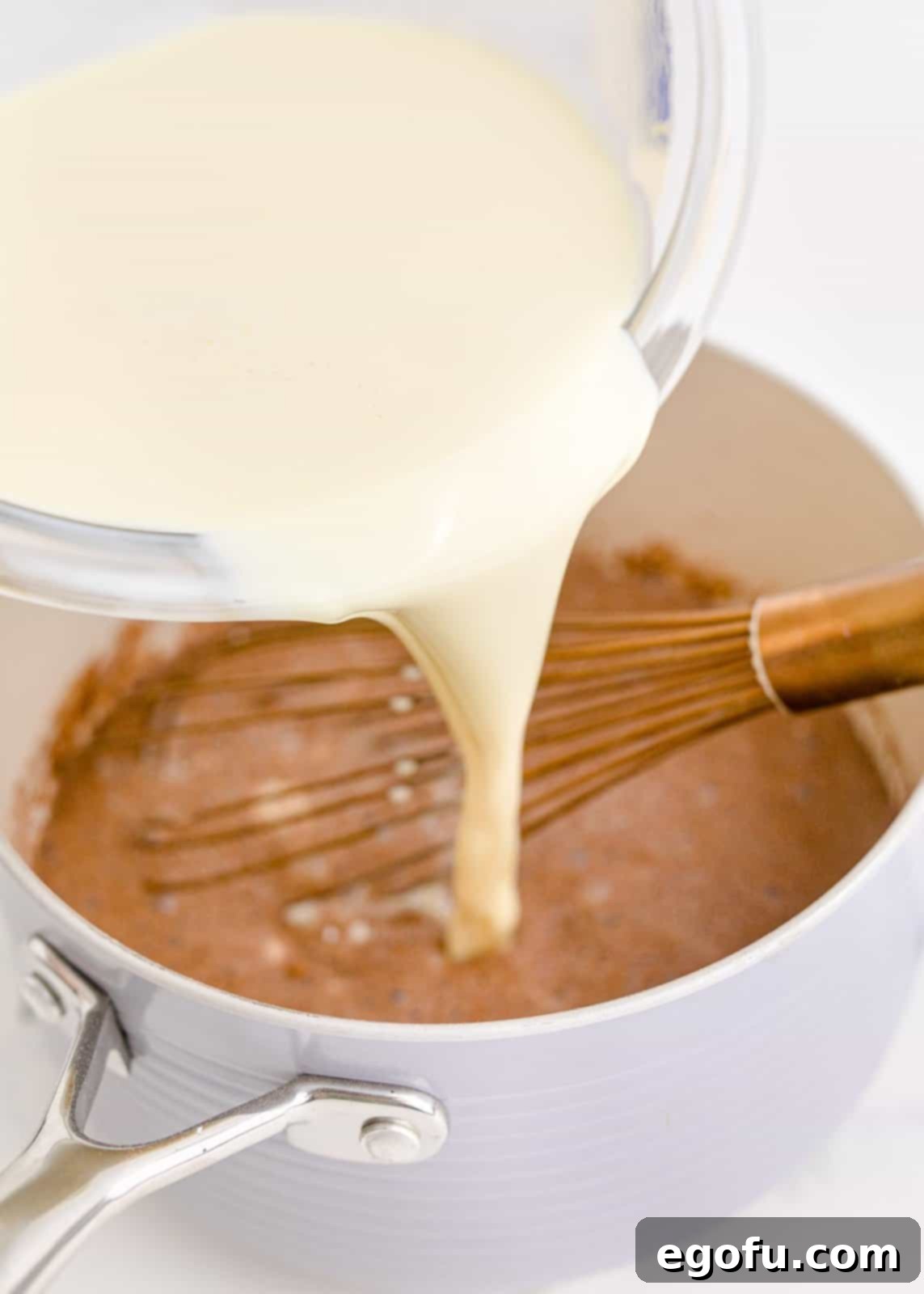Egg and half and half mixture slowly being added to the cocoa and sugar mixture in the pan, with a whisk in action.