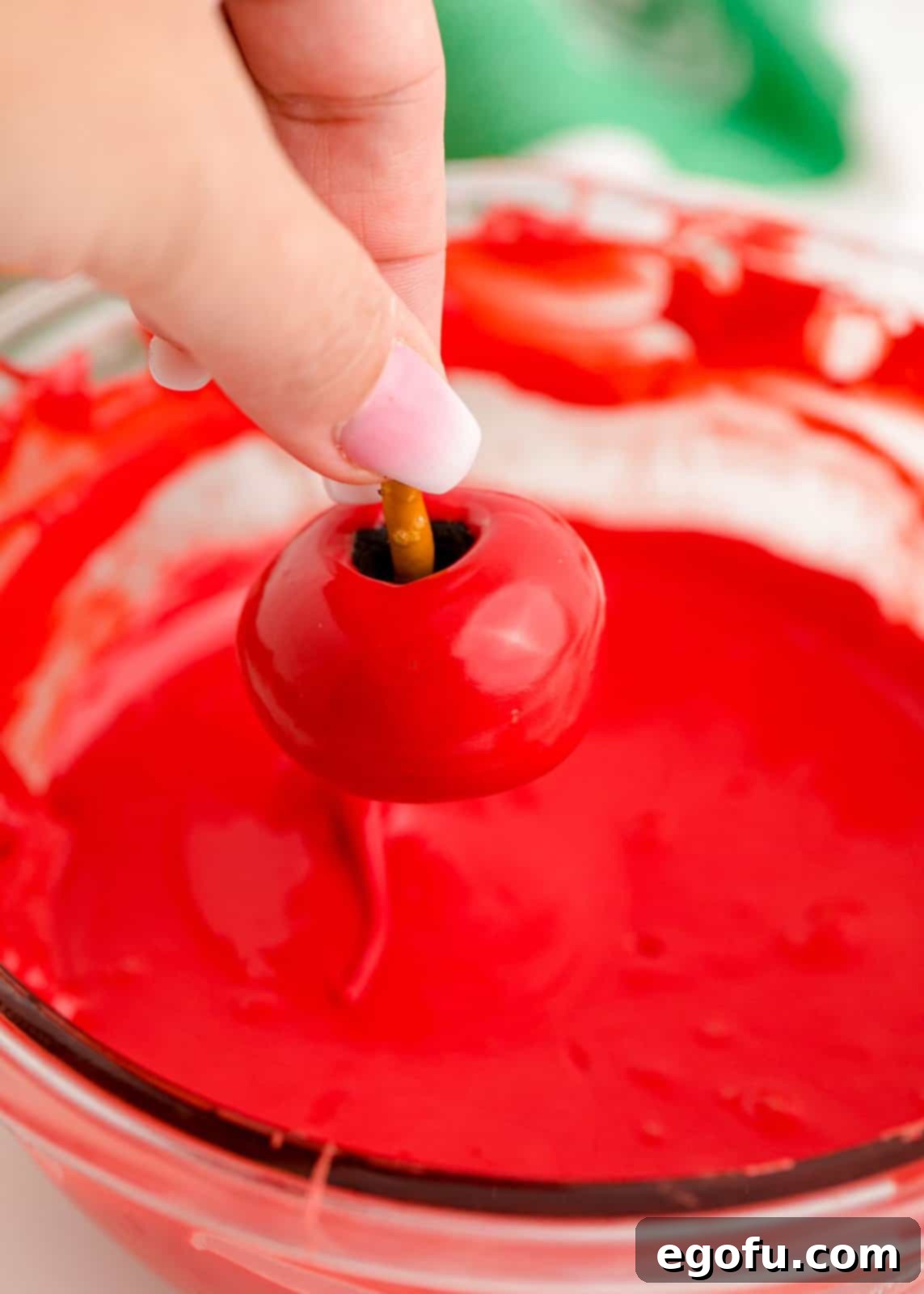 A hand dipping an apple-shaped Oreo truffle into a bowl of melted red chocolate, ensuring an even and complete coating for a vibrant finish.