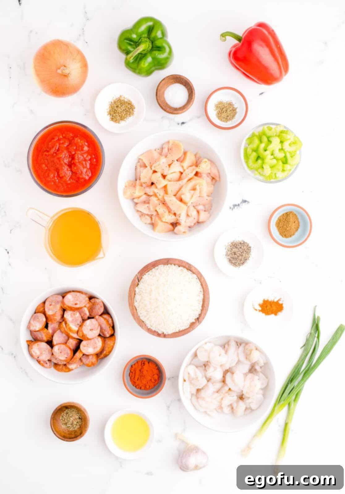 Assortment of fresh ingredients for Jambalaya including cooking oil, Andouille sausage, chicken, various bell peppers, celery, garlic, crushed tomatoes, and a selection of spices.