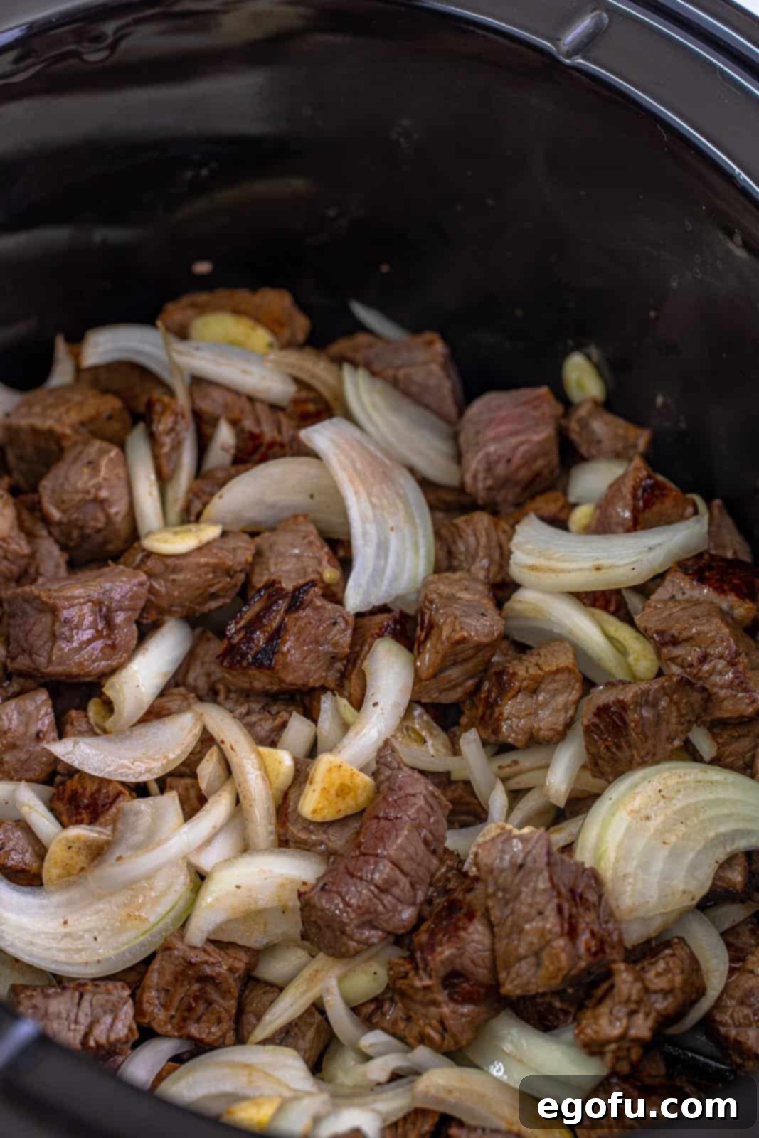 Onions and garlic added to the slow cooker with seared steak bites, ready for liquid.
