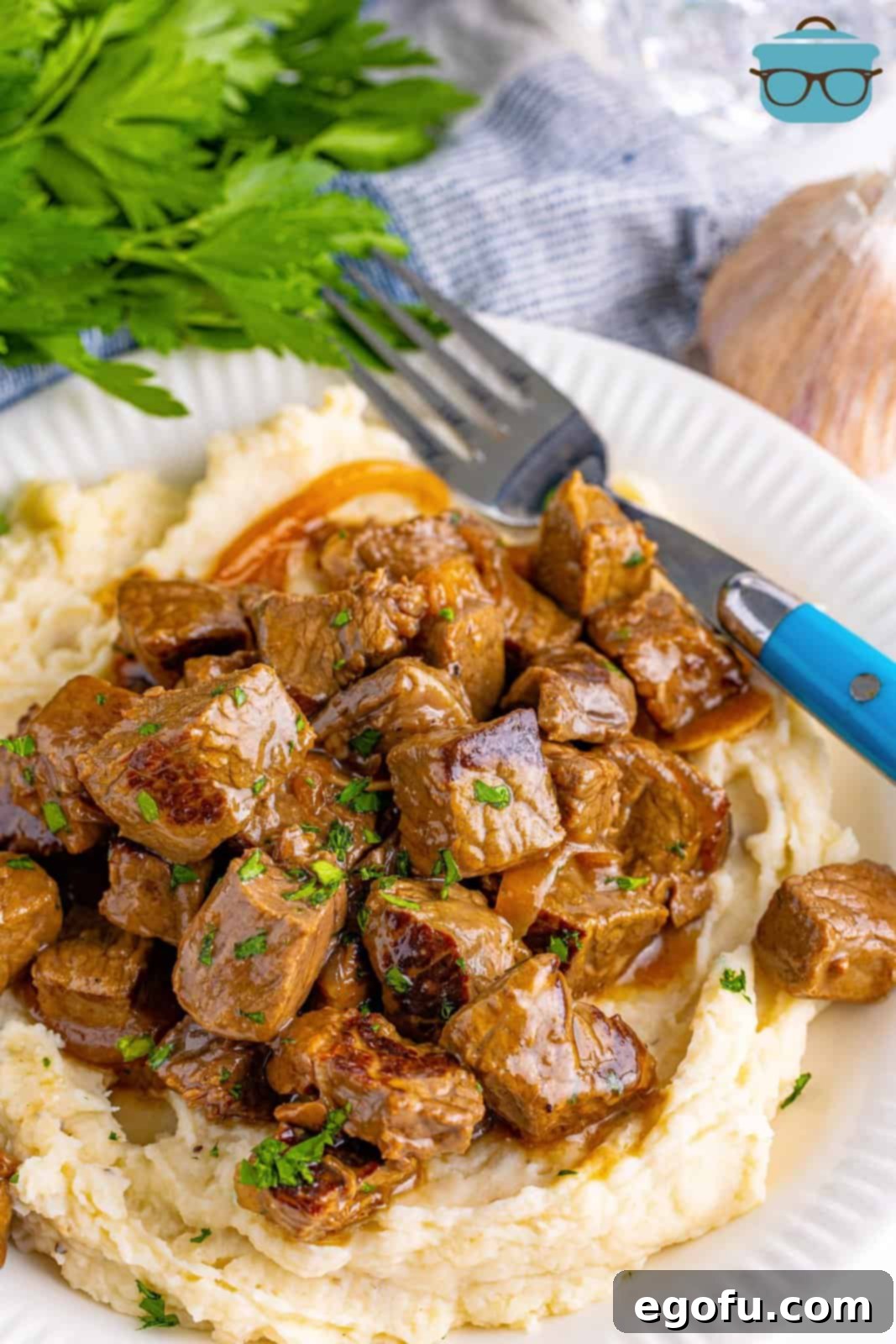 Overhead photo of Slow Cooker Garlic Butter Steak Bites over mashed potatoes on a white plate with a fork, ready to be eaten.