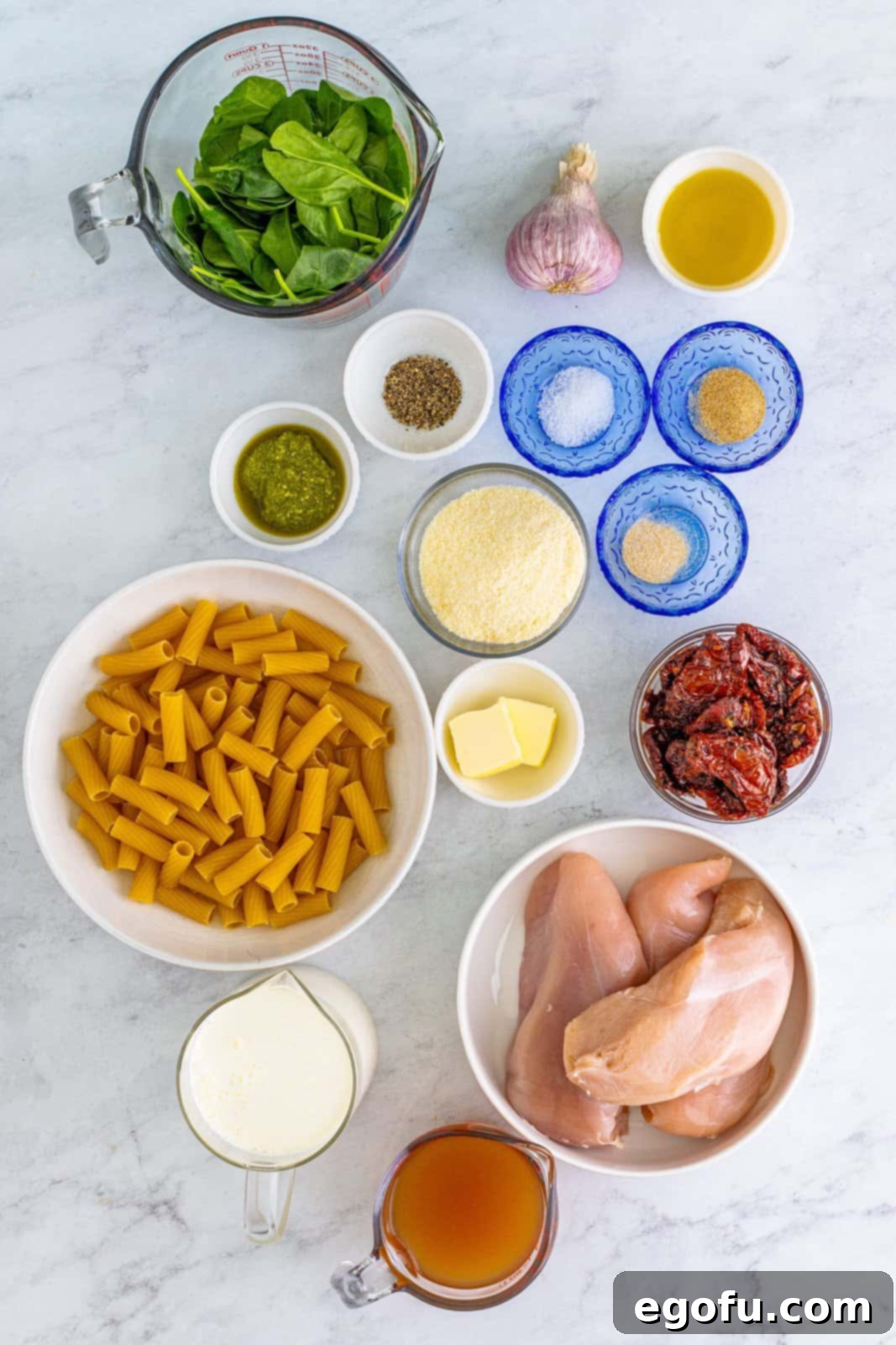 Ingredients needed: boneless skinless chicken breasts, kosher salt, pepper, garlic powder, onion powder, olive oil, salted butter, canned sun-dried tomatoes, garlic, prepared pesto, unsalted chicken stock, heavy cream, baby spinach, parmesan cheese and rigatoni or other short pasta, arranged on a counter.