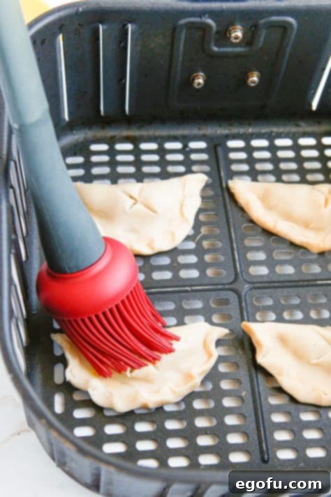 Uncooked hand pies in air fryer basket being brushed with egg wash.