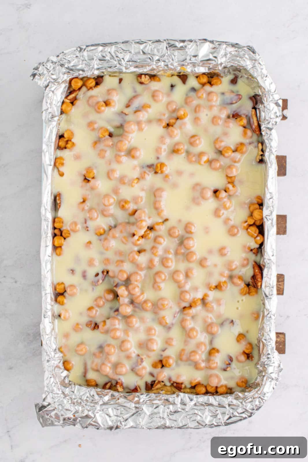 Sweetened condensed milk being poured evenly over the layers of chocolate chips, pecans, and caramel bits in the baking dish.