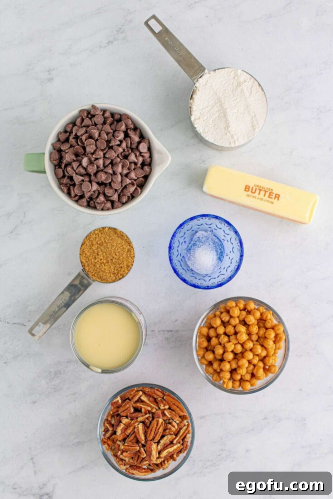 A beautiful flat lay of all the essential ingredients needed for Turtle Magic Bars: softened salted butter, packed light brown sugar, all-purpose flour, semi-sweet chocolate chips, chopped pecans, caramel bits, and a can of sweetened condensed milk.
