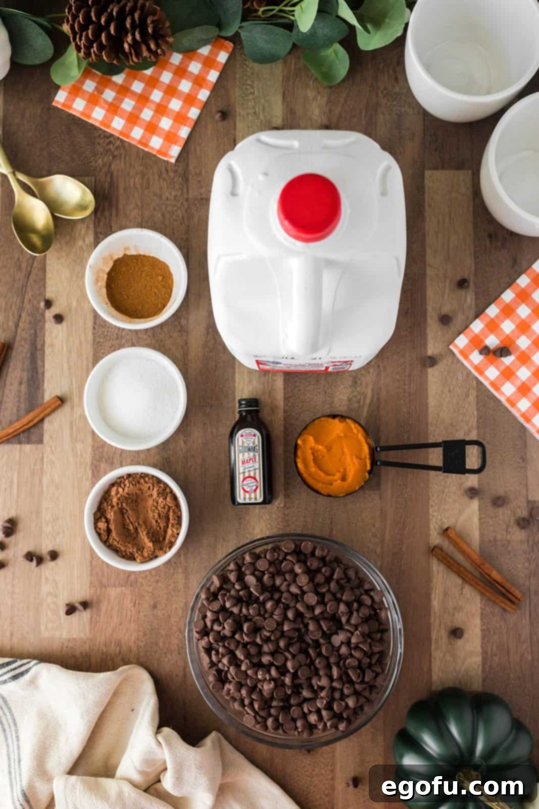 A flat lay photograph showcasing all the key ingredients needed for Crock Pot Pumpkin Hot Chocolate: a carton of whole milk, a can of pumpkin puree, a bowl of unsweetened cocoa powder, granulated sugar, milk chocolate chips, and pumpkin pie spice. Optional toppings like marshmallows, whipped cream, and chocolate sauce are also displayed.
