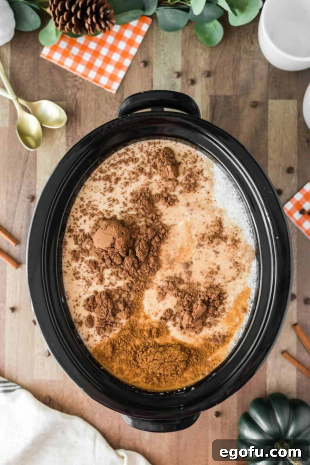 All the raw ingredients for pumpkin hot chocolate, including milk, pumpkin puree, cocoa, sugar, chocolate chips, and pumpkin pie spice, have been added to a crock pot and are being stirred together.