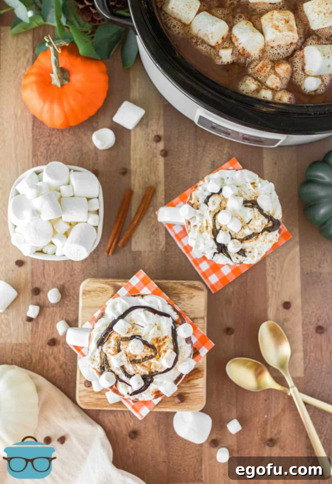 An overhead shot showcasing two mugs of Crock Pot Pumpkin Hot Chocolate, beautifully garnished with whipped cream, chocolate syrup, and cinnamon, placed on a rustic wooden table.