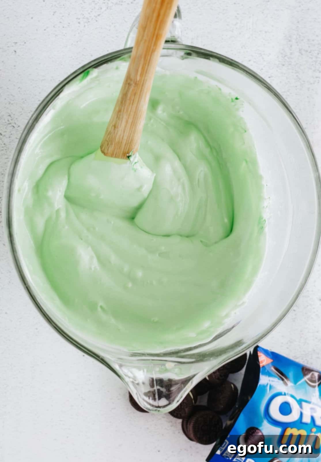 Cool Whip and green food coloring being carefully folded into the light green pudding mixture in a bowl.
